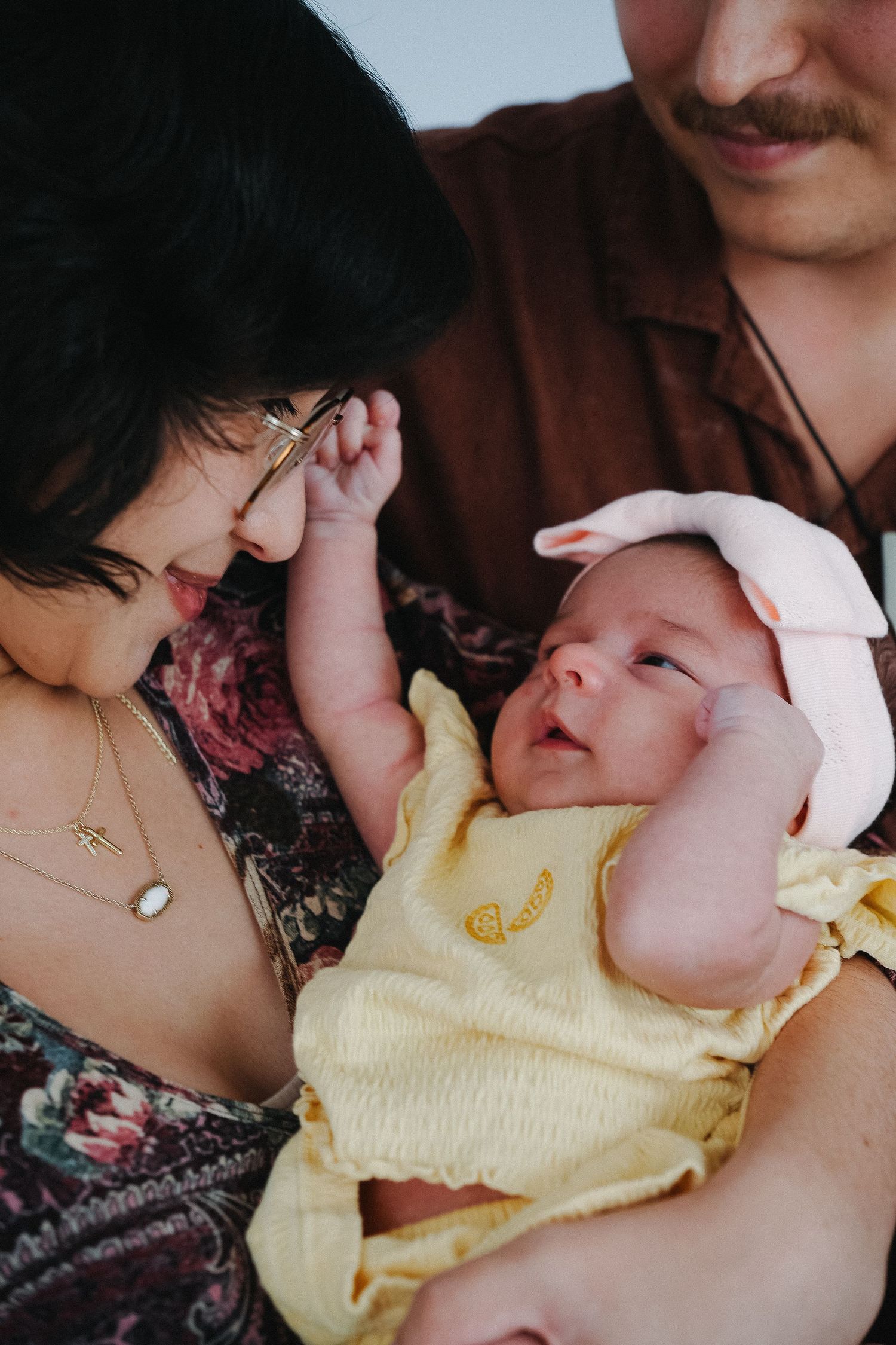 Parents lovingly holding their newborn baby dressed in yellow outfit with pink hat.