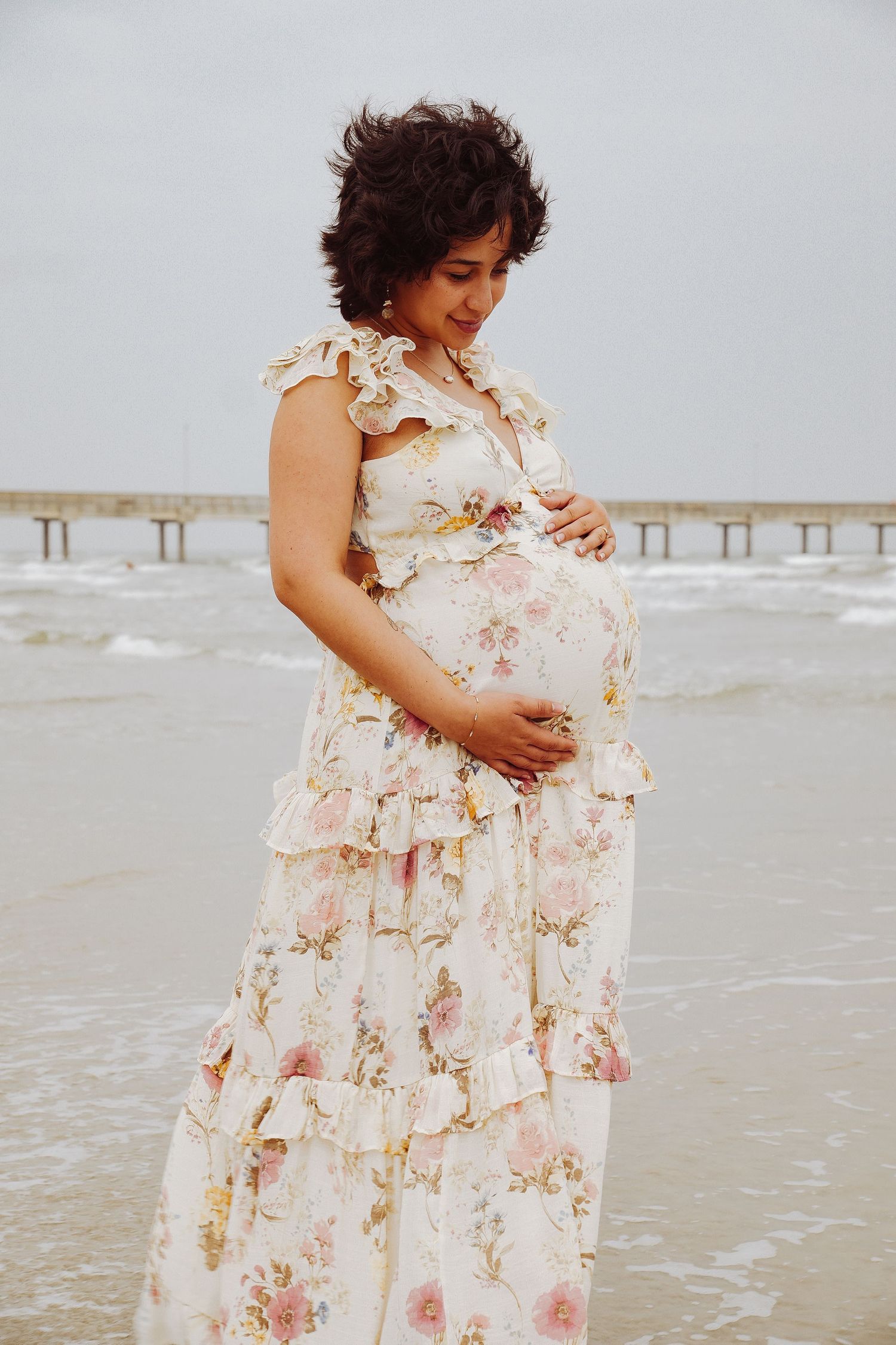 Pregnant woman in floral ruffle maxi dress cradling baby bump while standing at ocean beach shoreline.