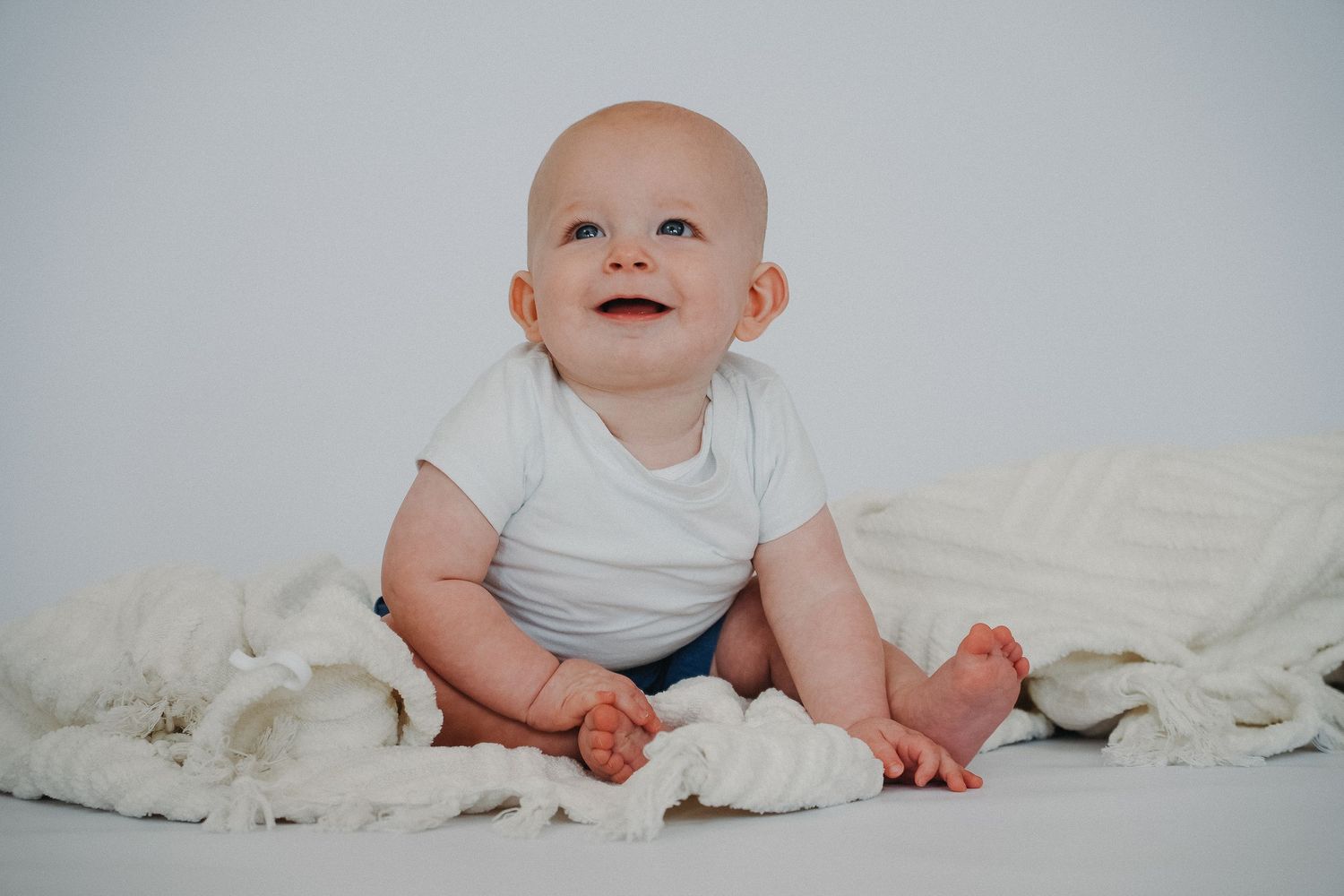 A happy baby in a white onesie sits smiling on a soft white blanket against a light background.