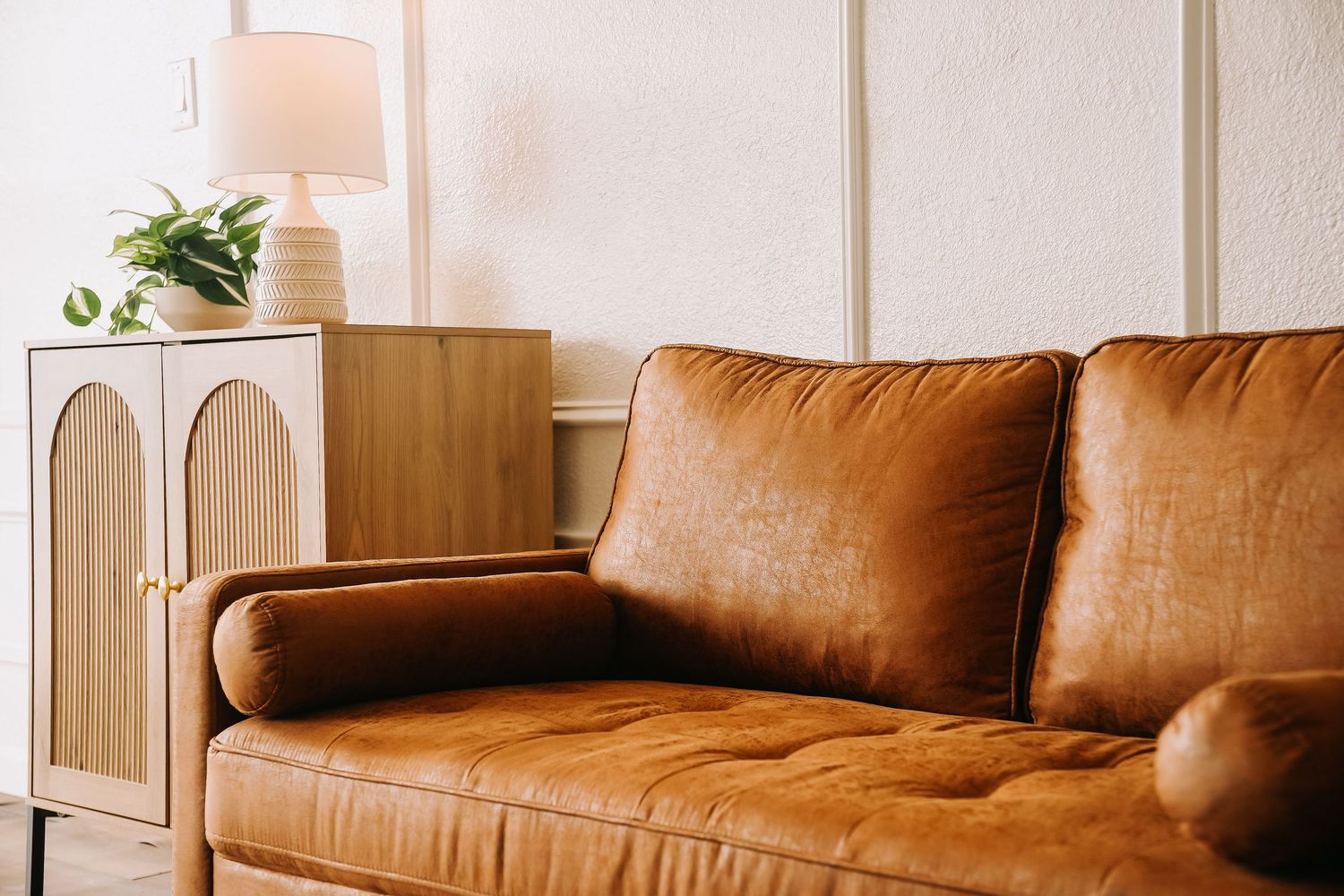 Cognac leather sofa beside a wooden arched cabinet with a white lamp in a bright, neutral living room.