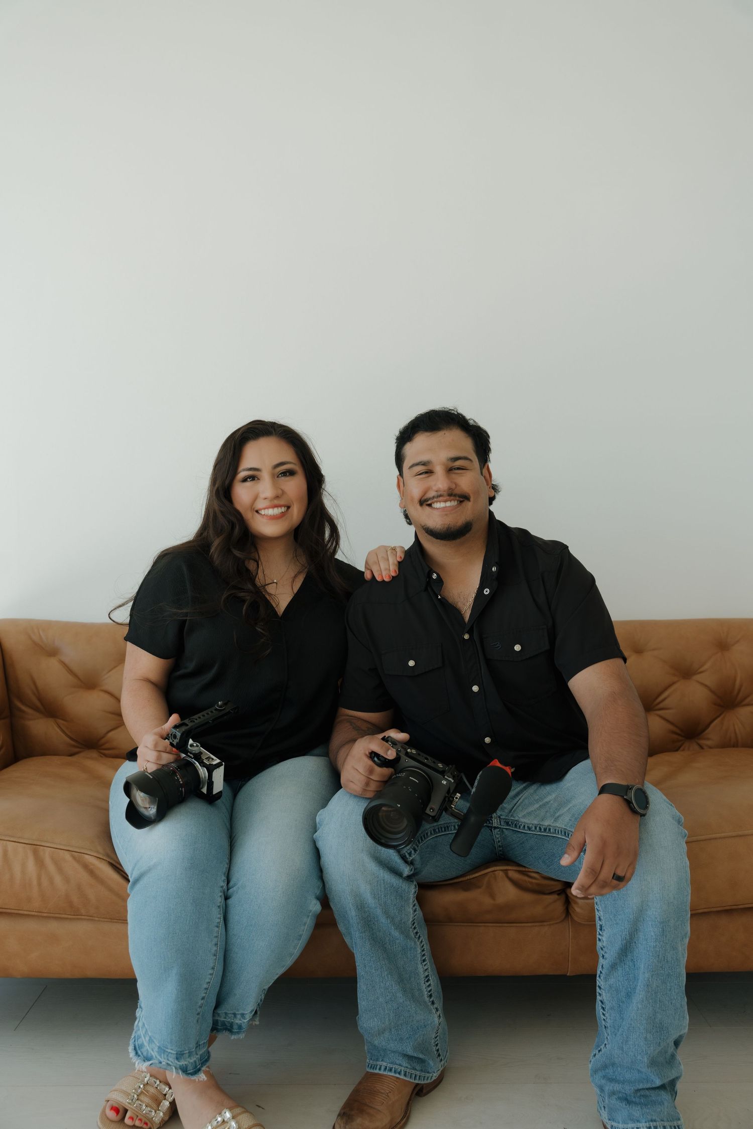 Smiling couple in black outfits sitting on leather sofa holding cameras.