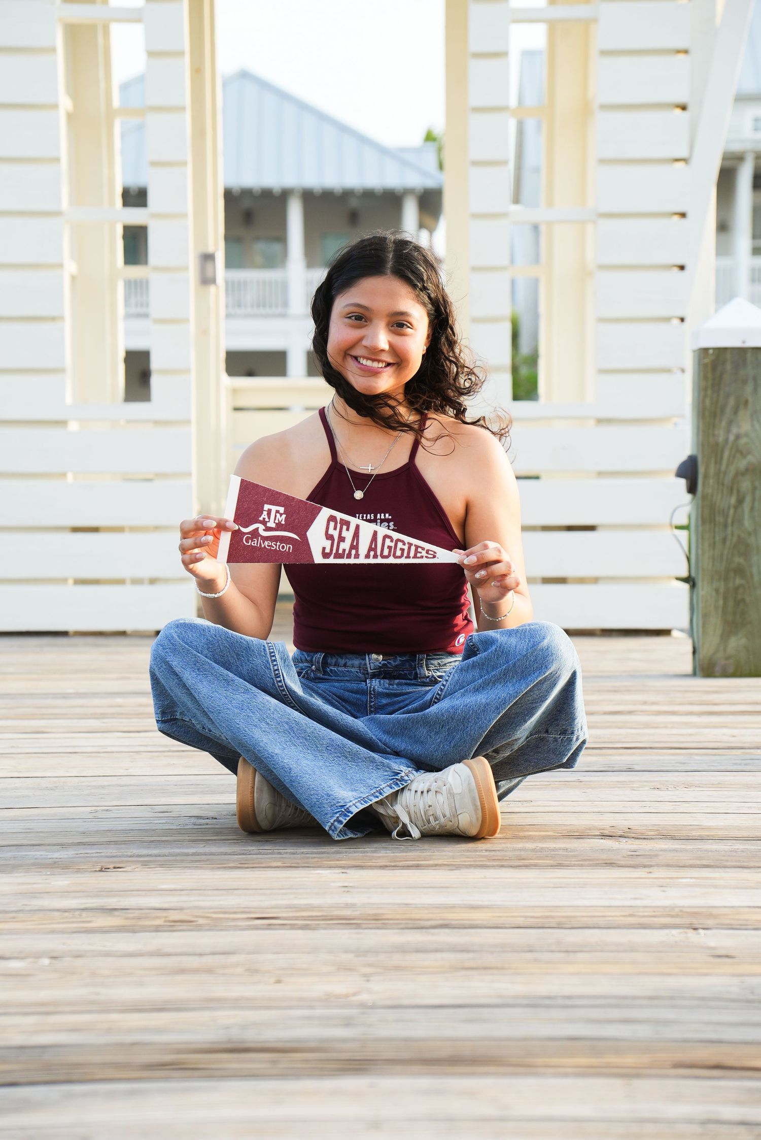 Smiling young woman sits cross-legged on wooden deck holding a Texas A&M Galveston Sea Aggies pennant.