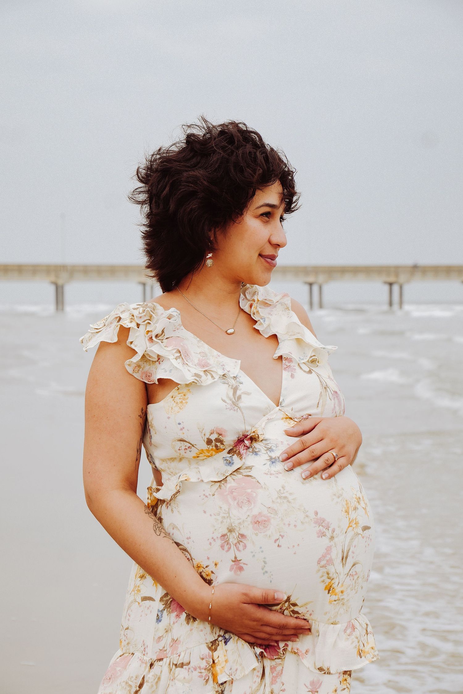 Smiling pregnant woman in floral dress holding baby bump at beach with ocean pier in background.