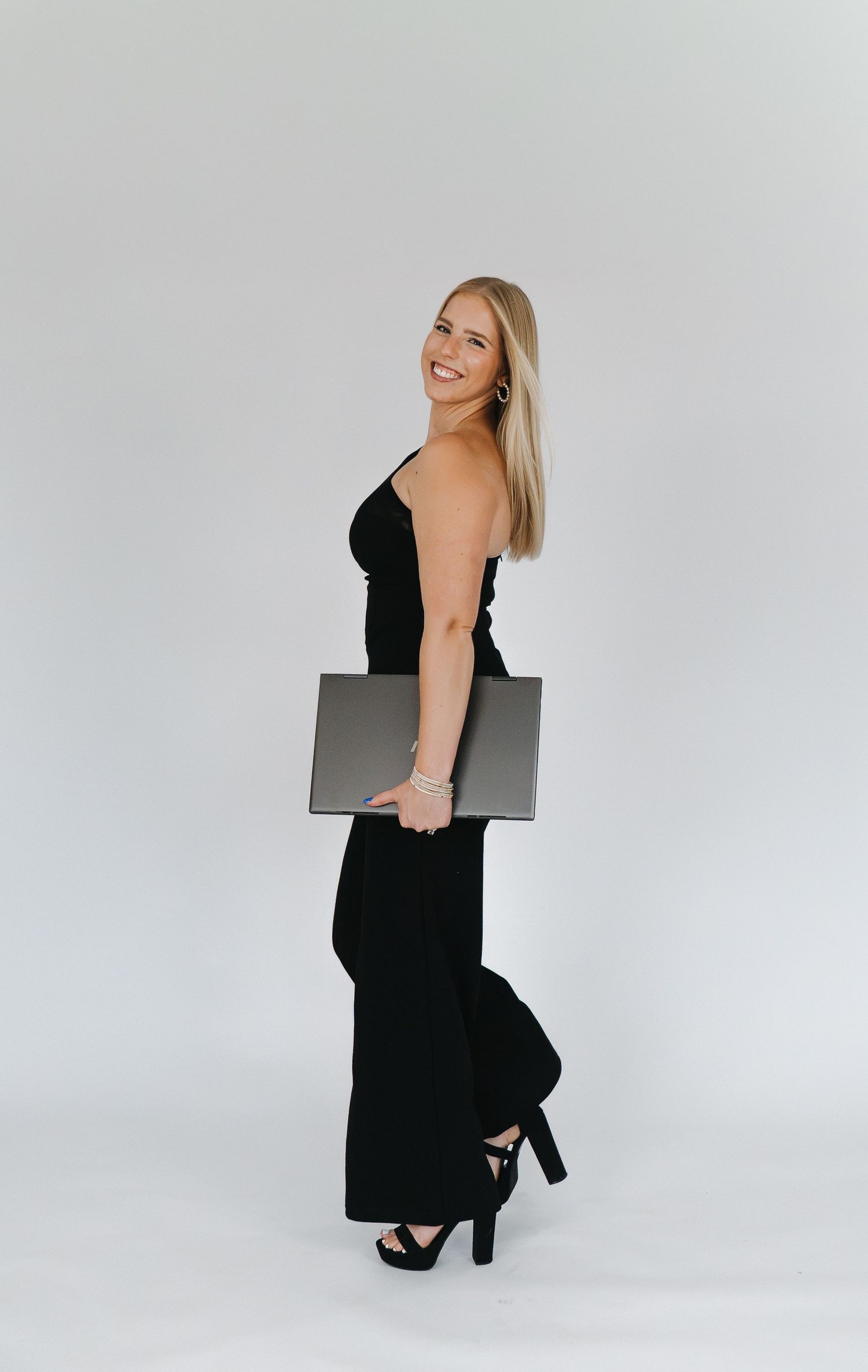 Smiling blonde woman in black jumpsuit and heels holding a laptop, posing against a white background.