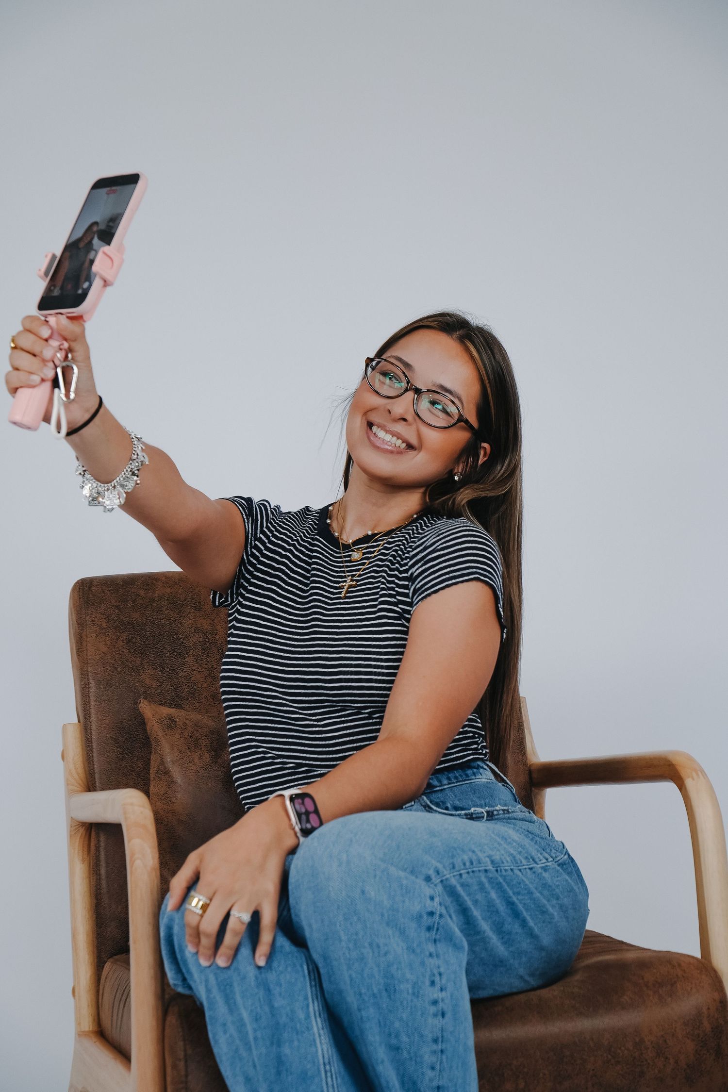 Smiling young woman in striped shirt and glasses taking a selfie while seated in a brown leather chair.