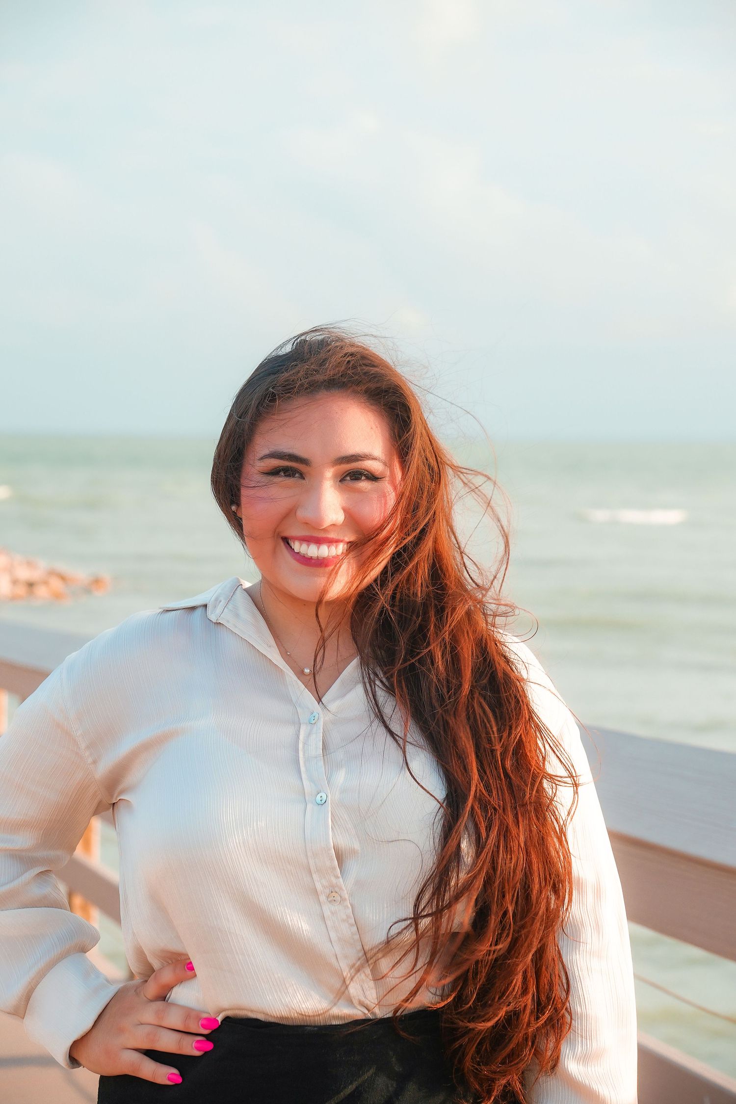 Smiling woman with long hair in white shirt poses by ocean breeze.