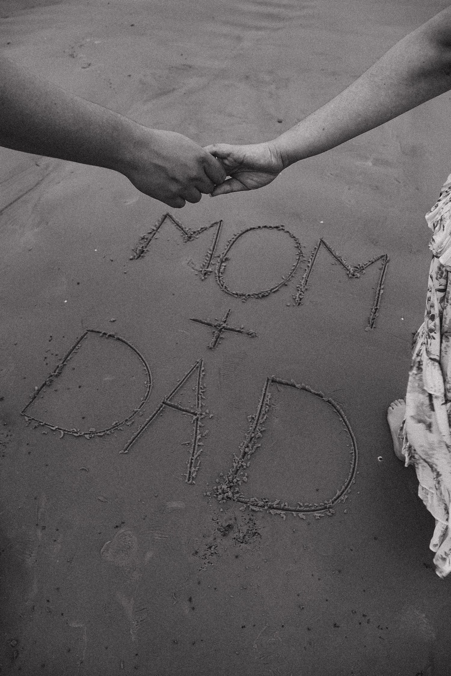 Black and white photo of a couple holding hands on a beach with MOM + DAD written in the sand.