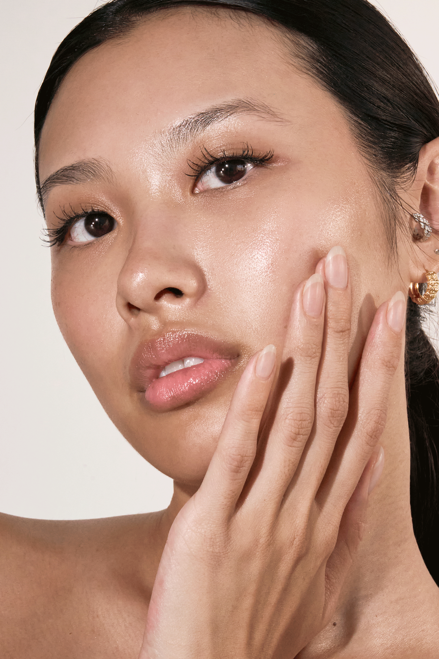 Close-up of a woman with glowing skin touching her cheek, wearing gold earrings against a neutral background.