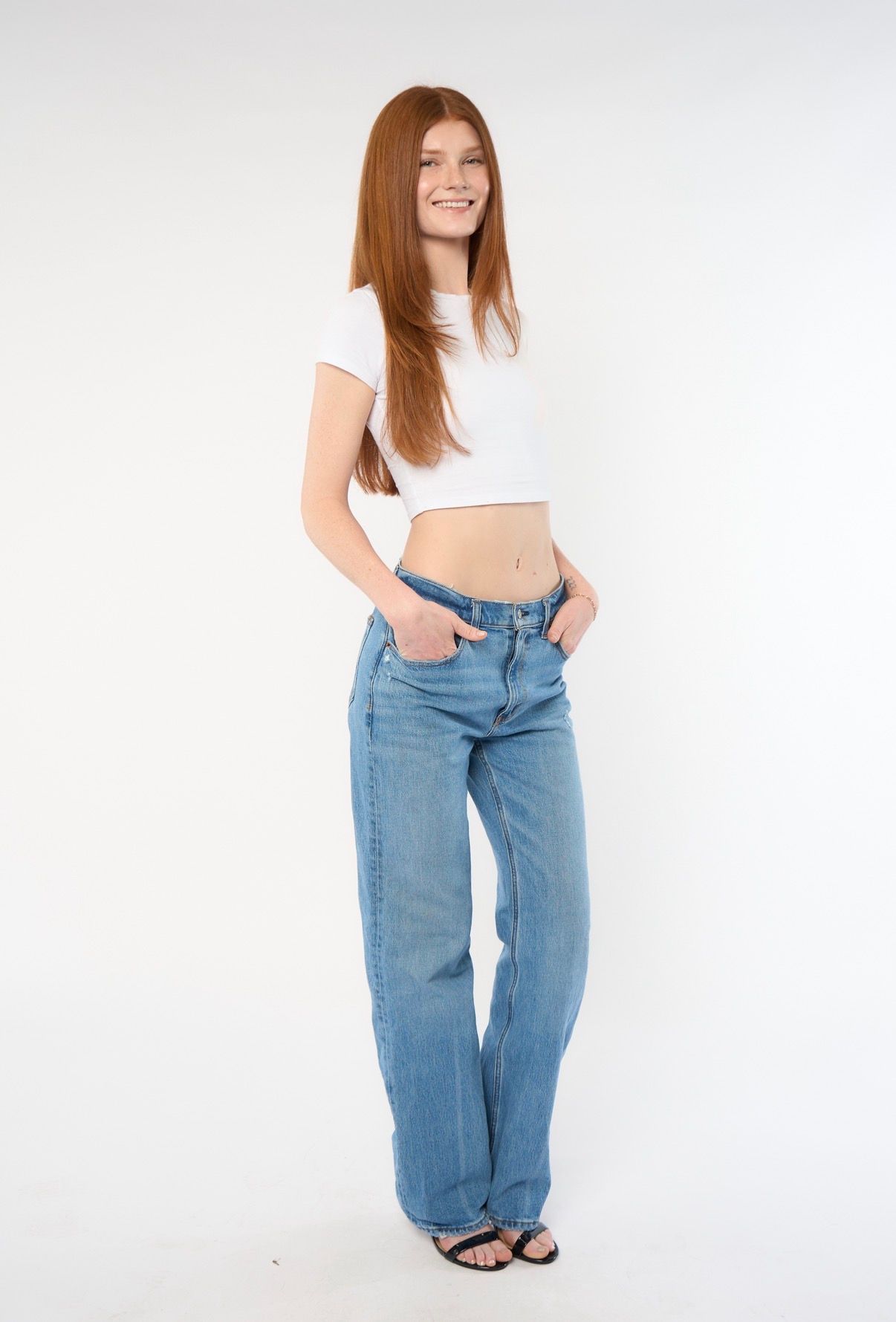 Young woman with red hair wearing a white crop top and light blue wide-leg jeans, smiling against a white background.