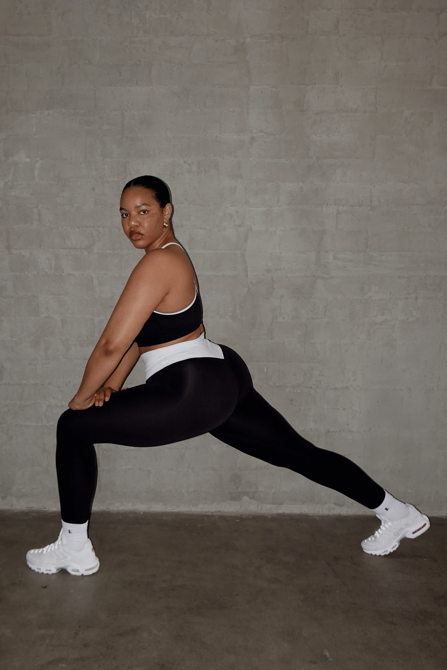Woman in black and white sports bra and leggings performing a lunge stretch against a gray concrete wall.