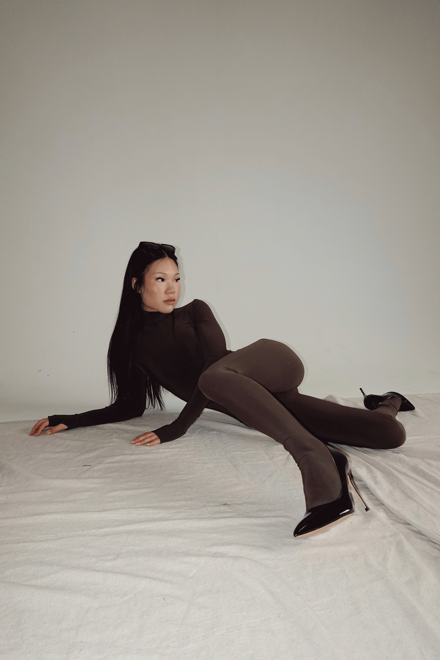 Woman in dark brown bodysuit and tights poses gracefully on white fabric in a studio setting.
