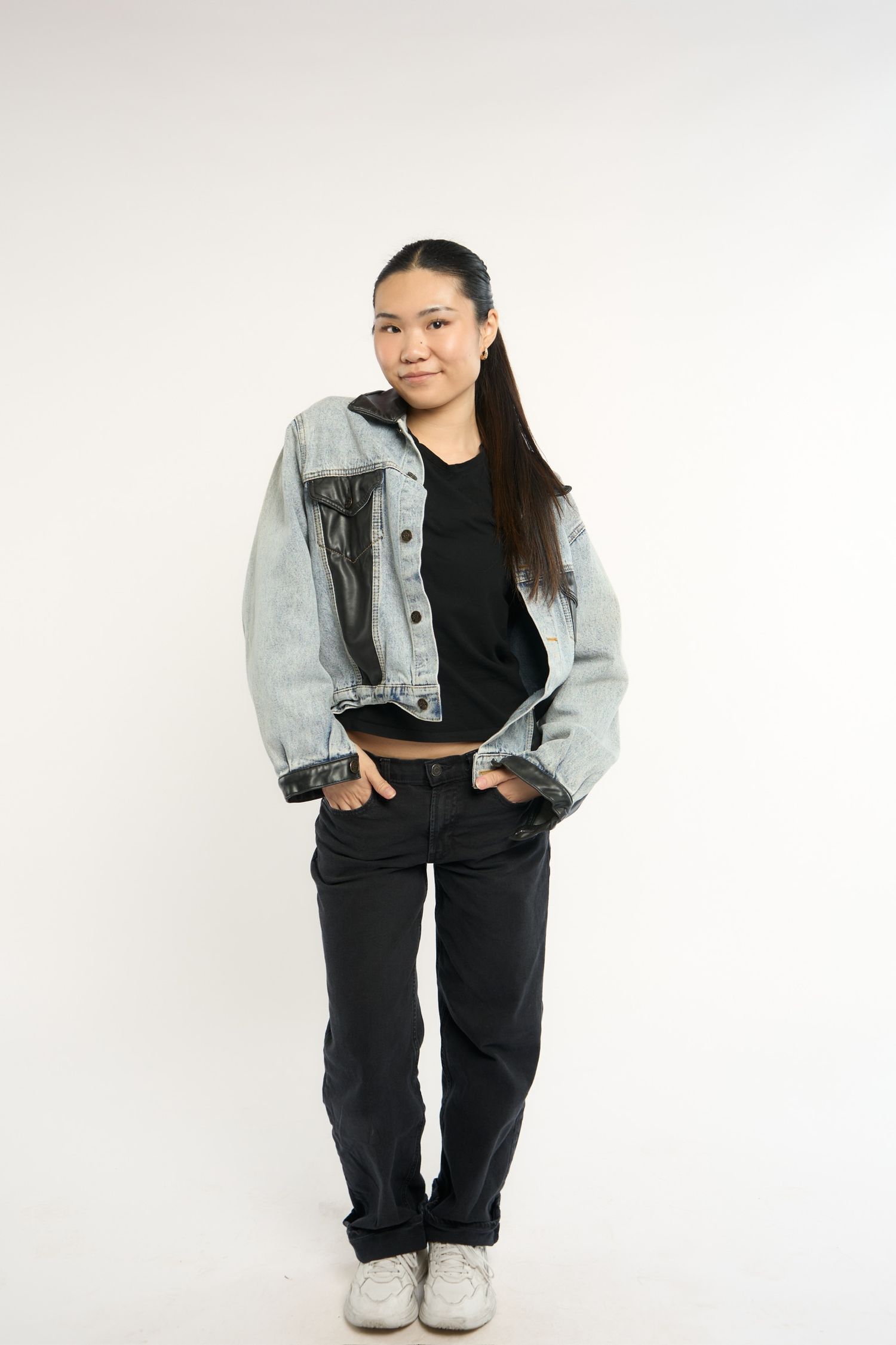 Young Asian woman in oversized denim jacket, black top and jeans, white sneakers, posing confidently on white background.