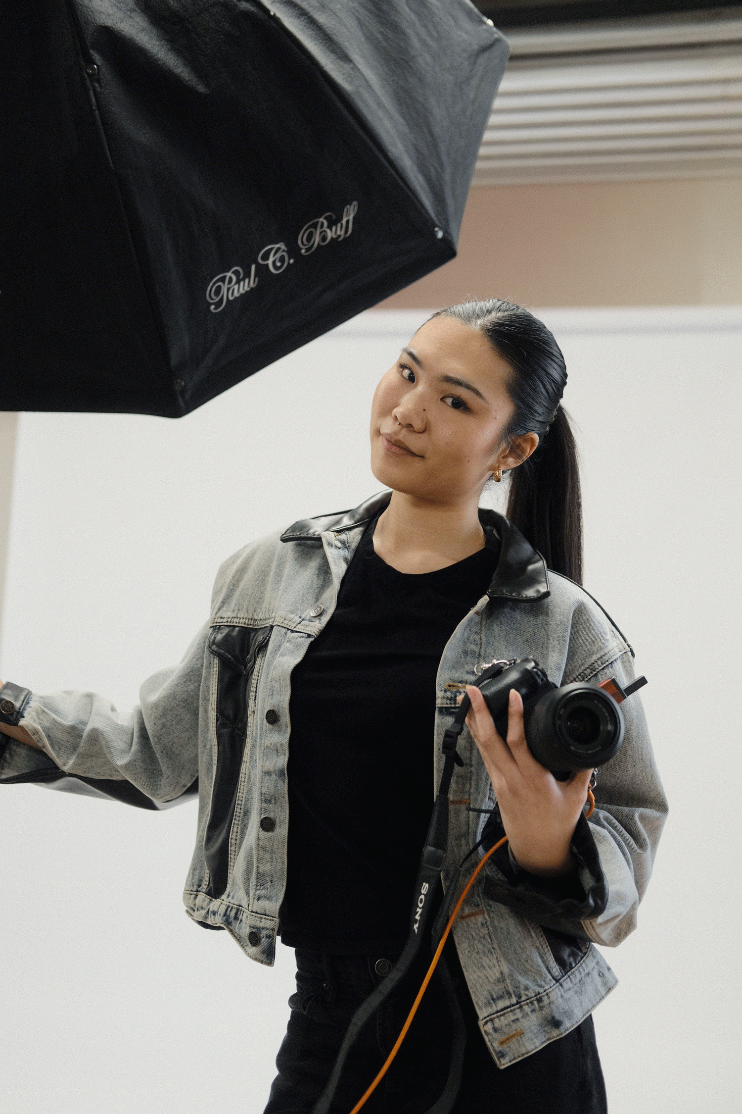 Young woman in denim jacket holding a camera and studio light umbrella in a photography studio.