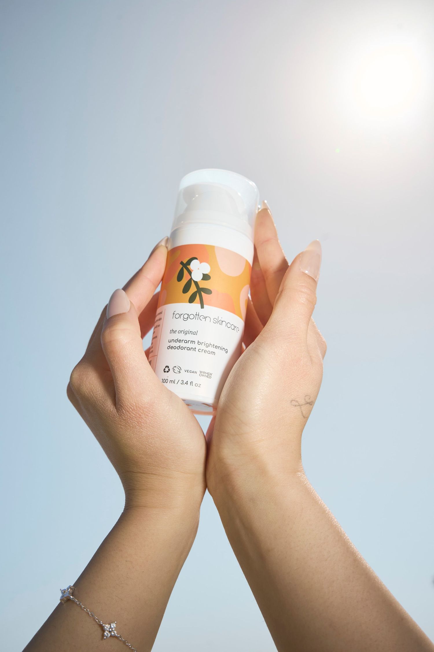 Hands holding Forgotten Skincare underarm brightening deodorant cream in orange packaging against sunny blue sky.