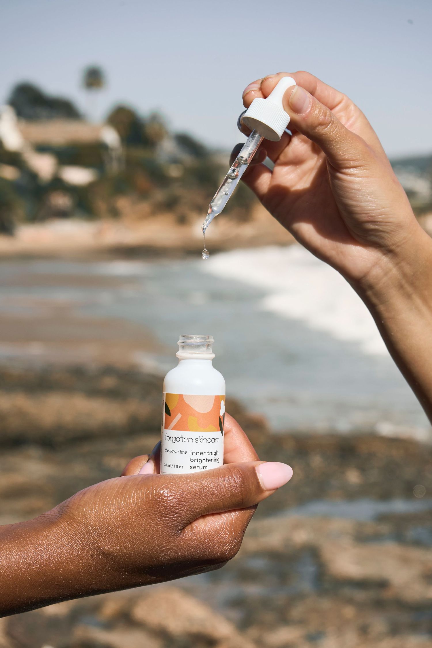 Hands holding a dropper and serum bottle from Forgotten Skincare at a rocky beach.