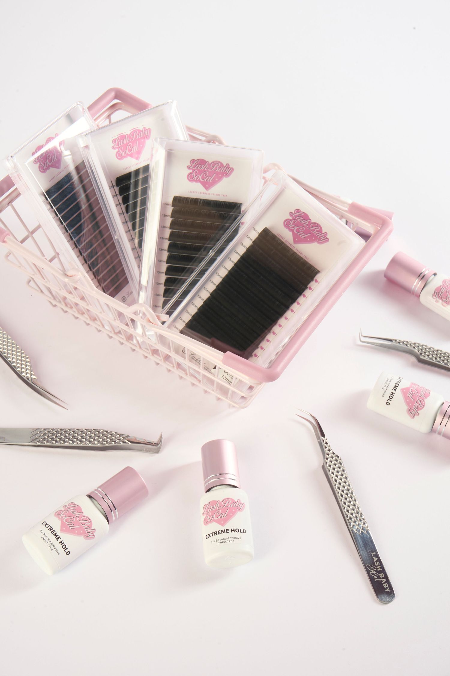Pink basket with eyelash extension trays, tweezers, and Extreme Hold adhesive bottles arranged on pink background.