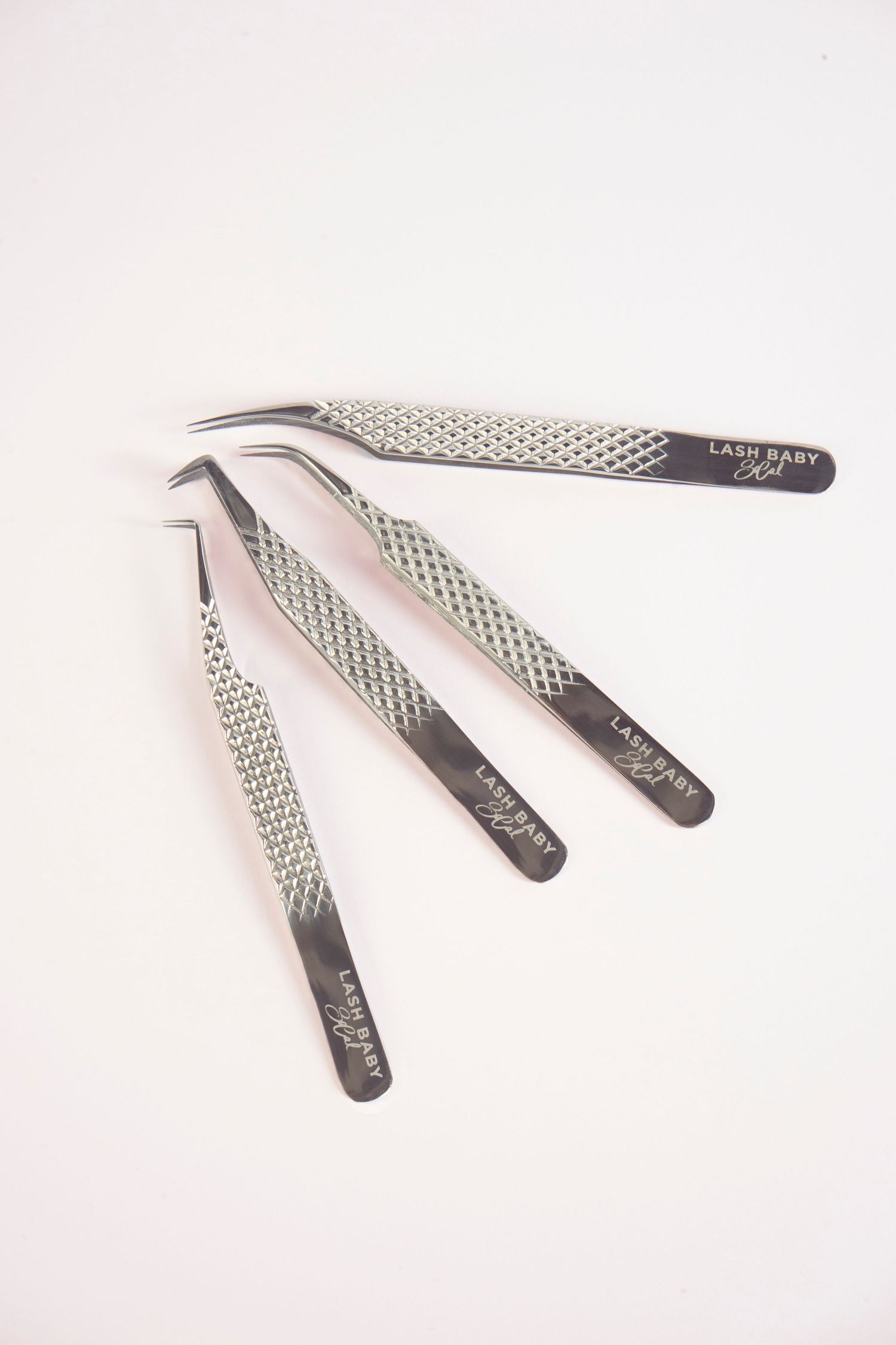 Four Lash Baby SoCal branded stainless steel tweezers with diamond-pattern grip handles on pink background.