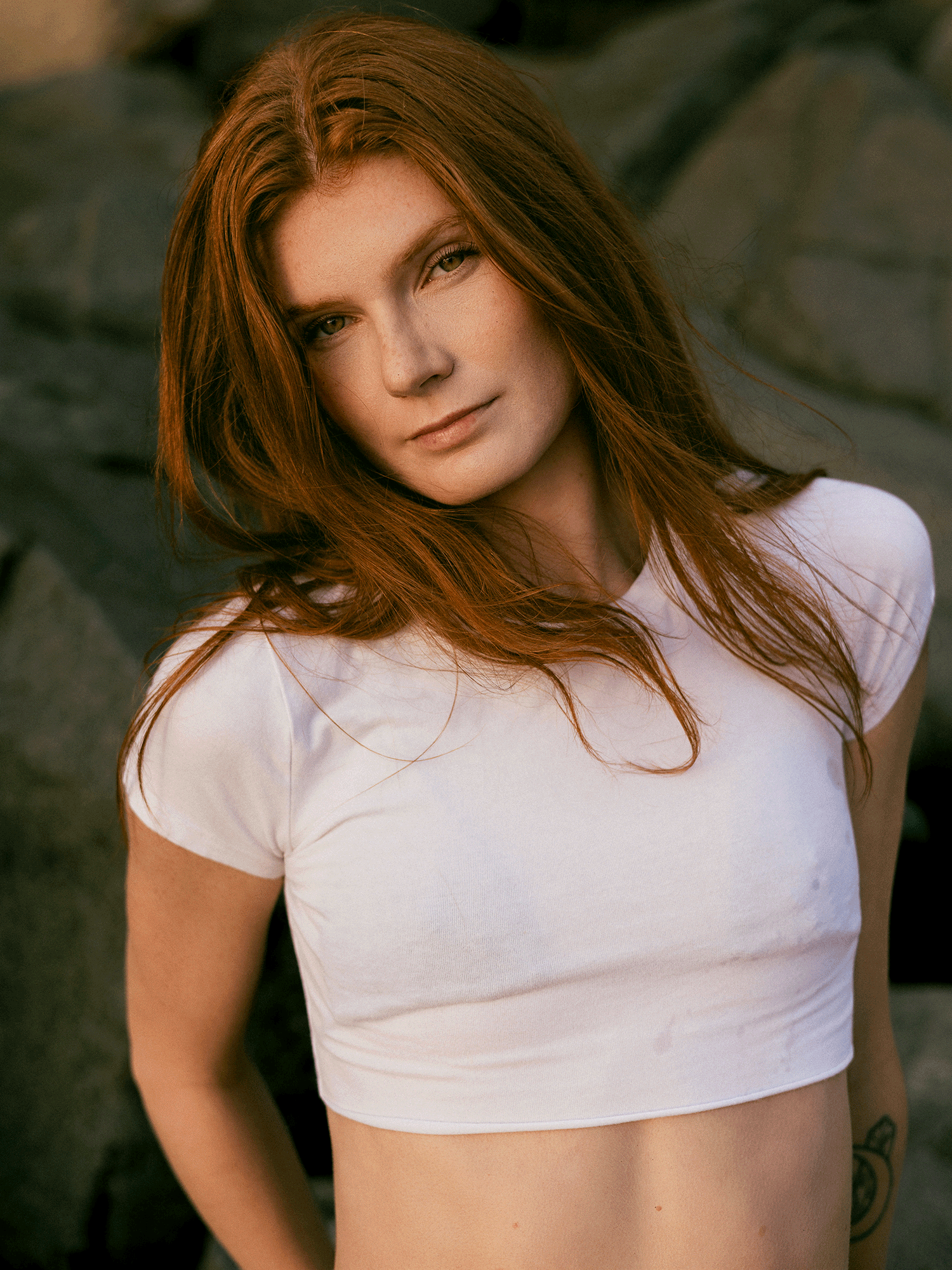 A young woman with long red hair wearing a white crop top poses confidently outdoors near rocky terrain.