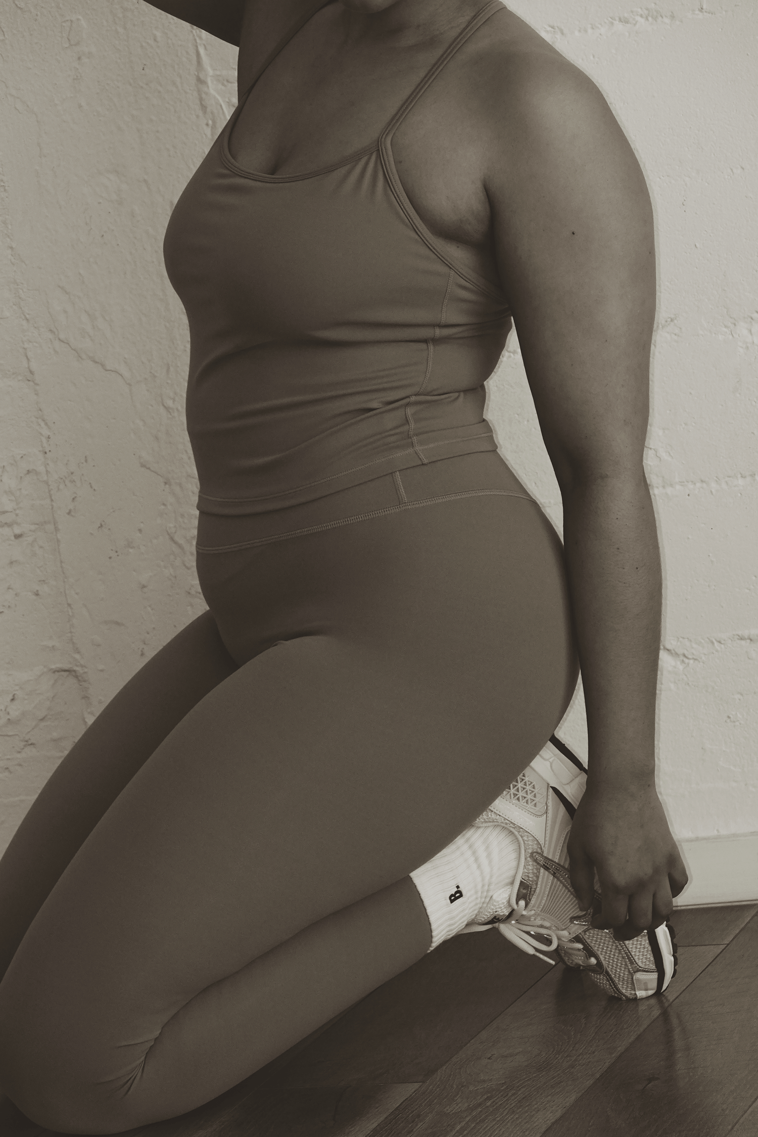 Woman in sports bra and high-waist leggings kneeling on floor holding white sneakers.
