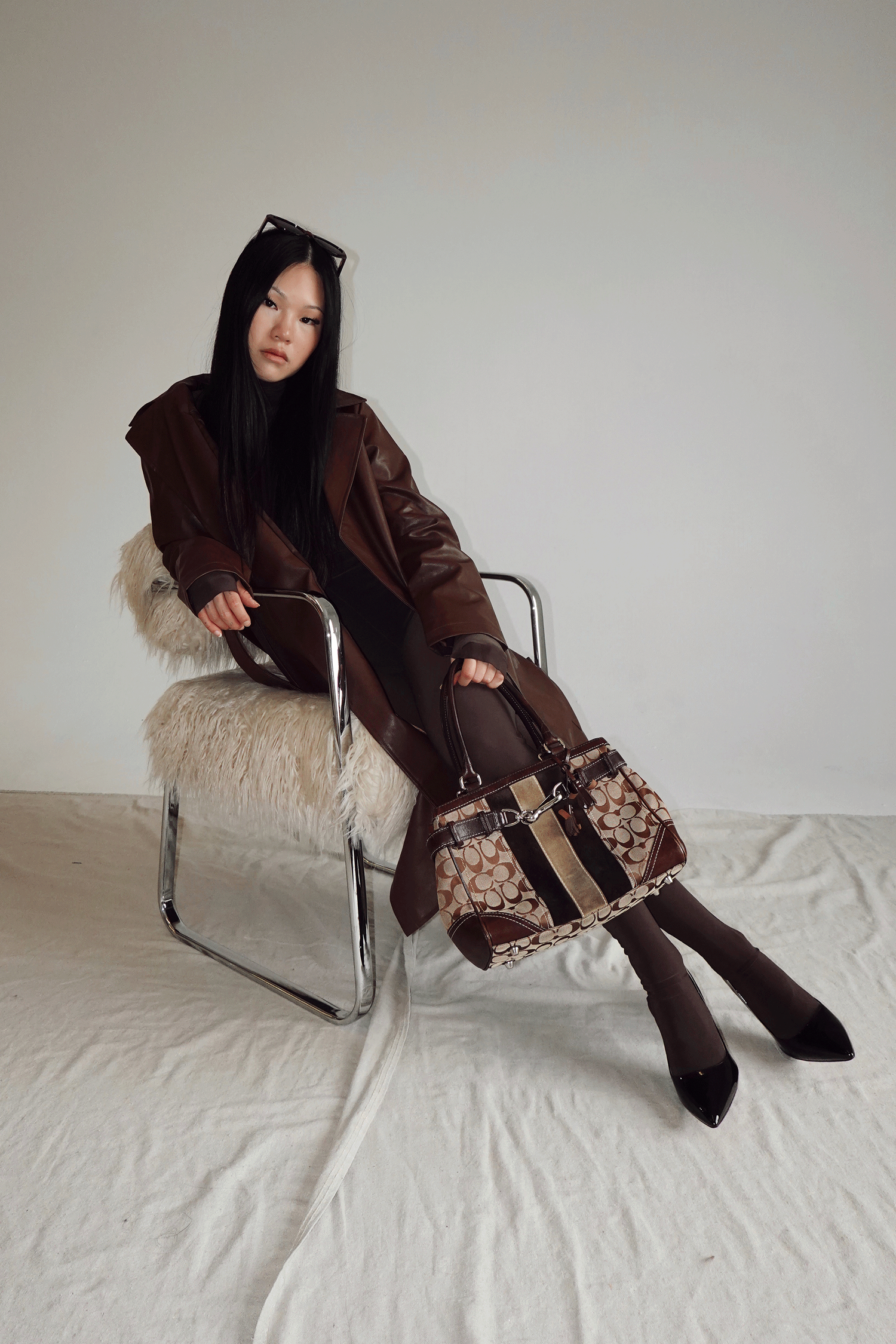 Woman in brown leather coat sits on fur chair holding a Coach monogram handbag, styled with dark tights and heels.
