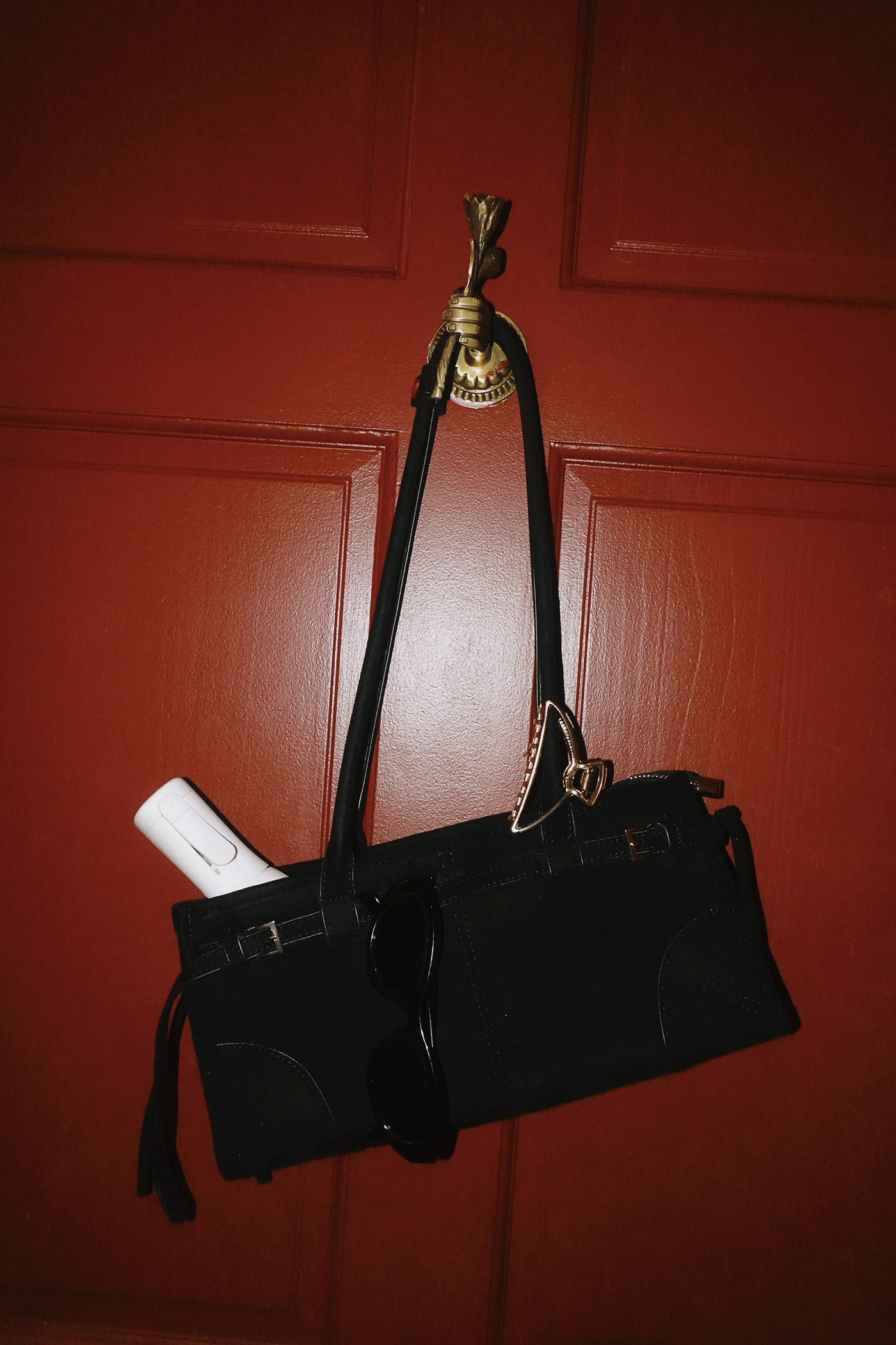 Black leather handbag with gold hardware hanging on a red door hook, with sunglasses and a white item inside.