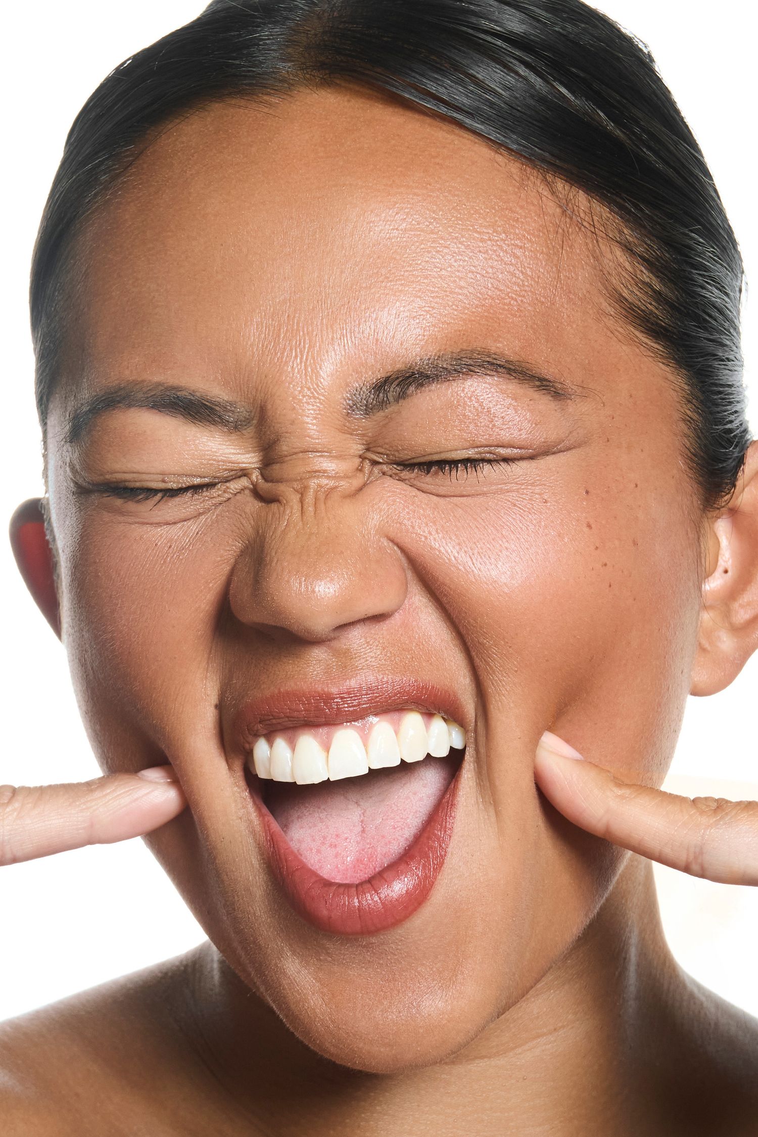 Young woman making an excited face with eyes closed, fingers pushing cheeks, mouth open wide showing white teeth.