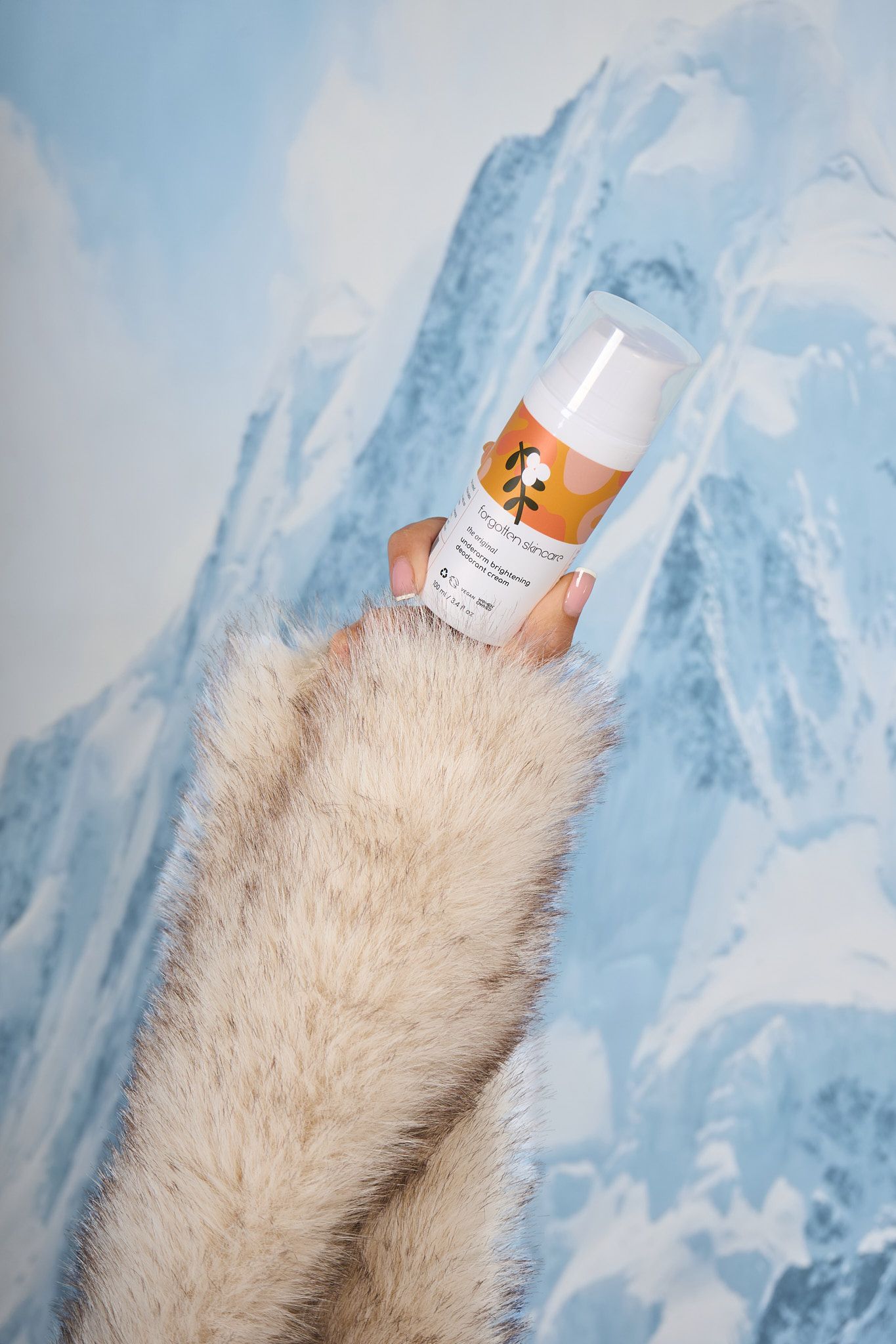 Hand in fur sleeve holding orange skincare product against snowy mountain backdrop.