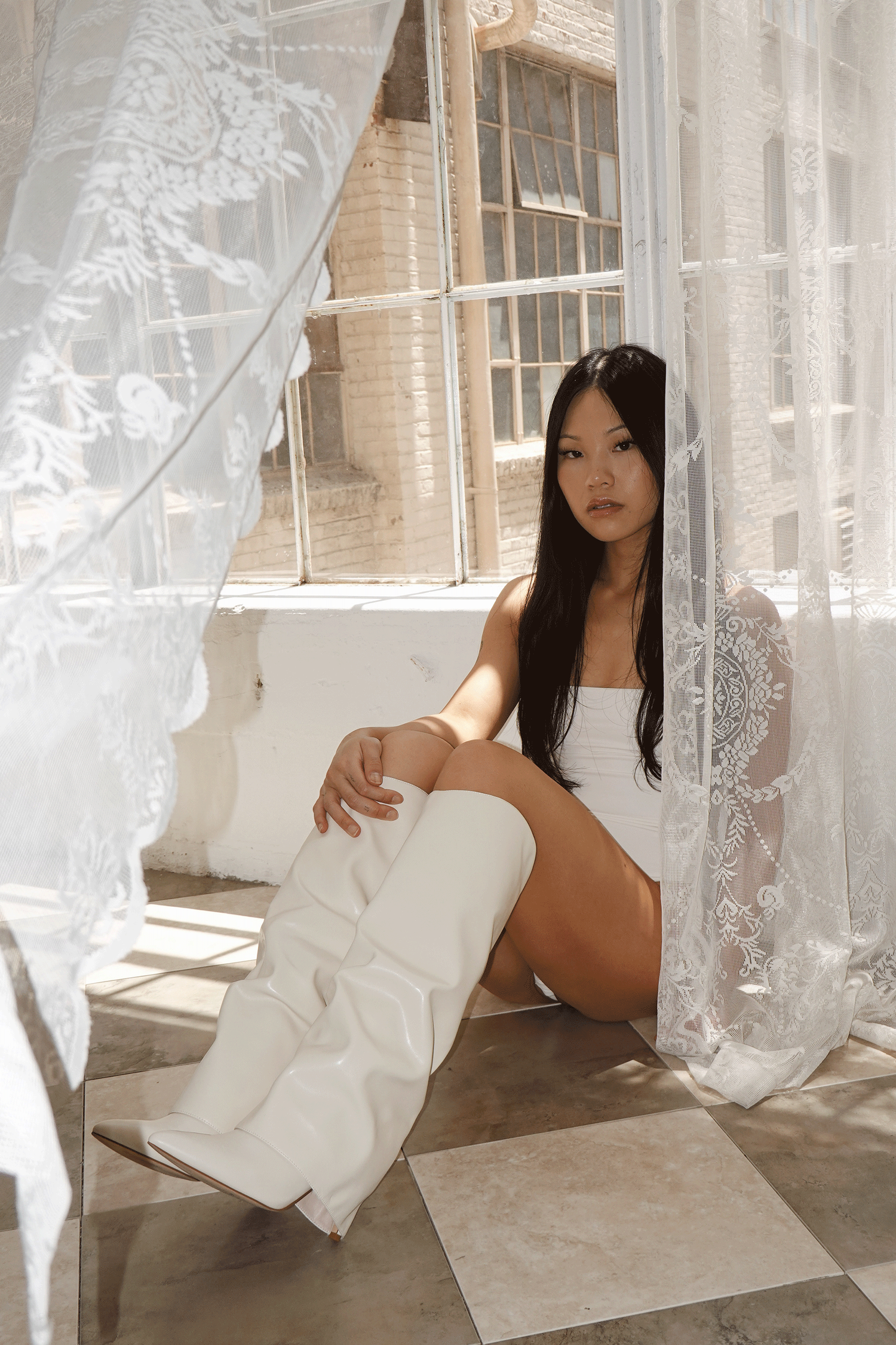 Young woman in white bodysuit and white boots sitting gracefully by sheer lace curtains near a sunlit window.