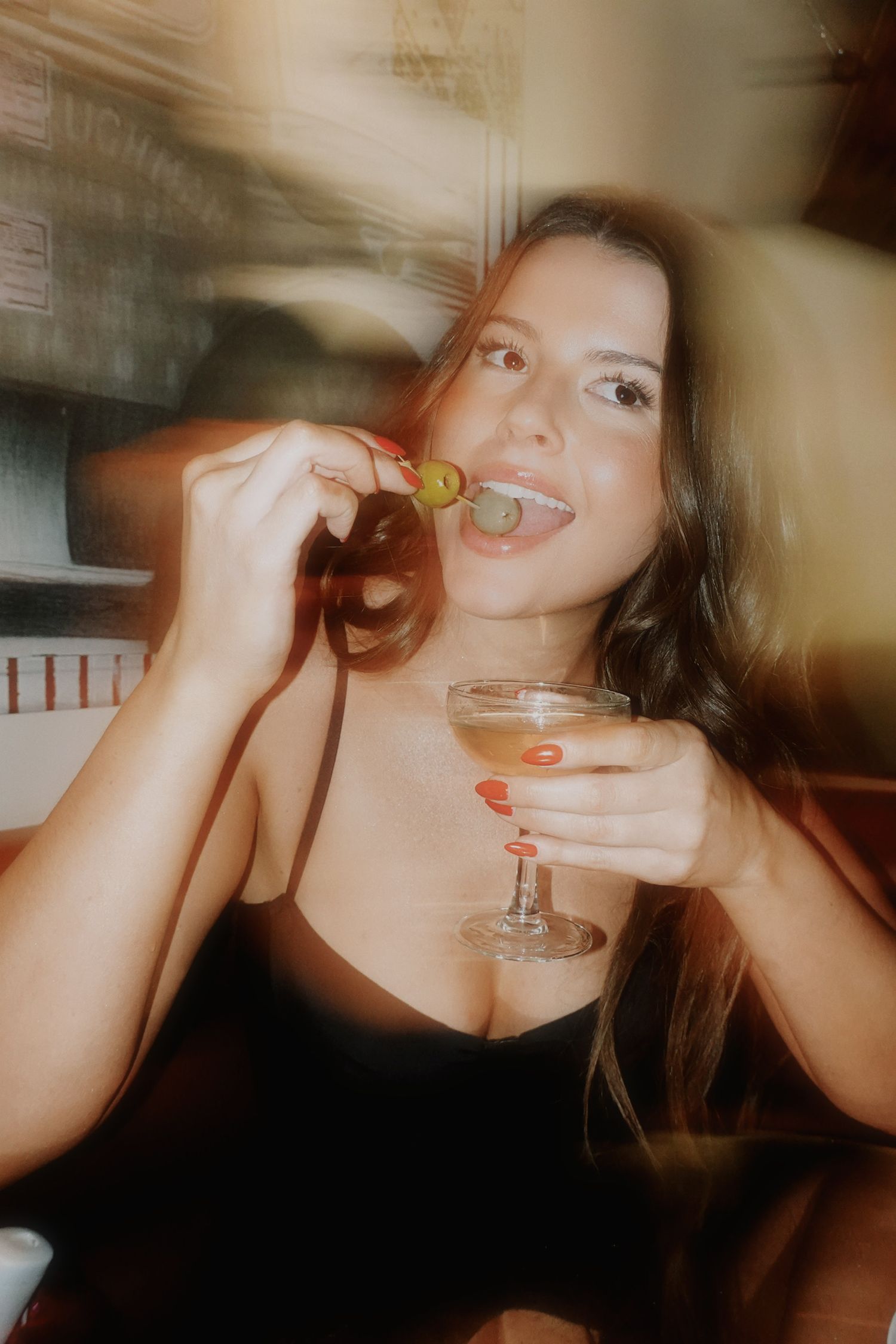 Woman in black dress enjoying a dirty martini with olive garnish at a bar, smiling with red nails.