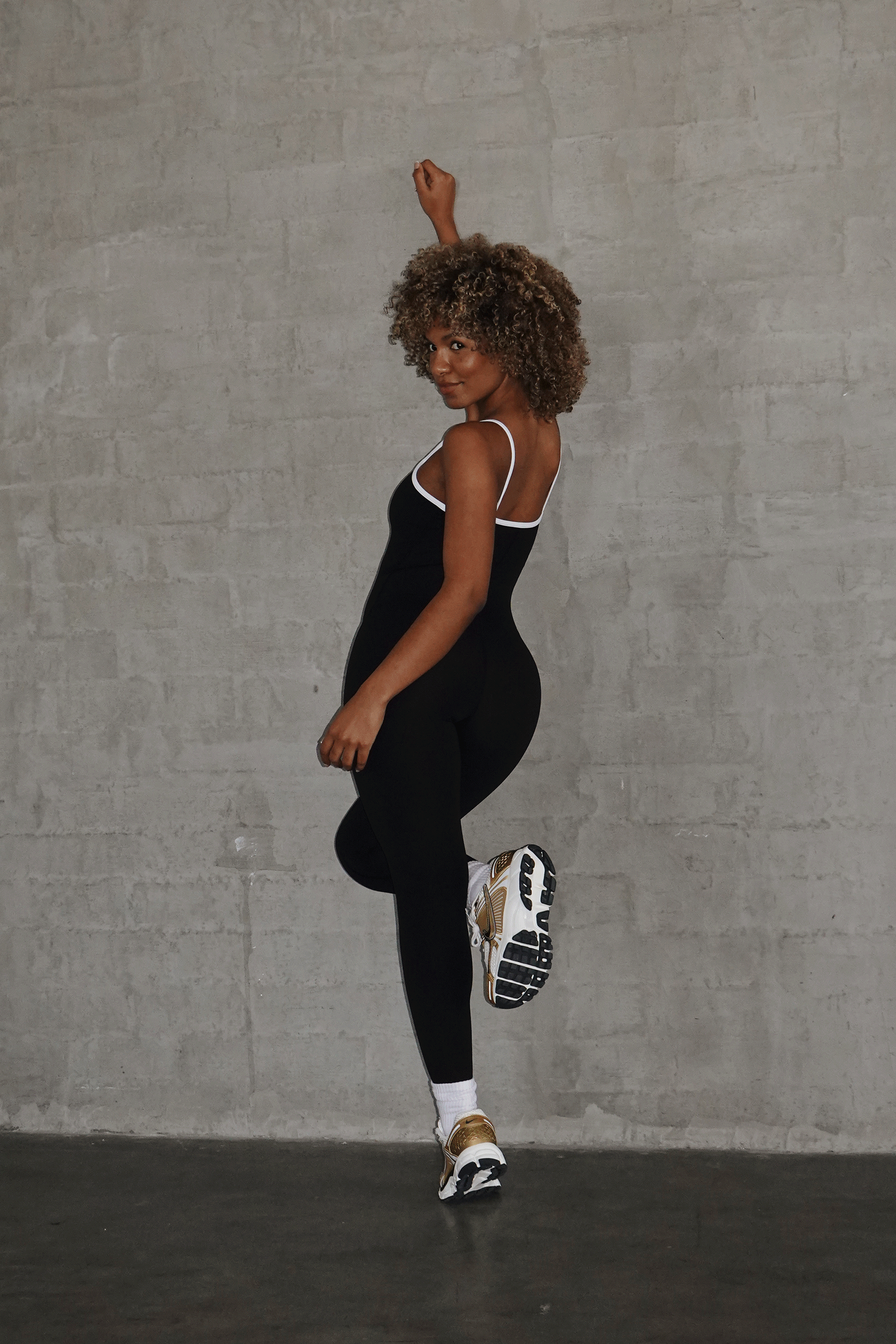 A woman in a black athletic jumpsuit and sneakers poses dynamically with one knee raised against a gray wall.