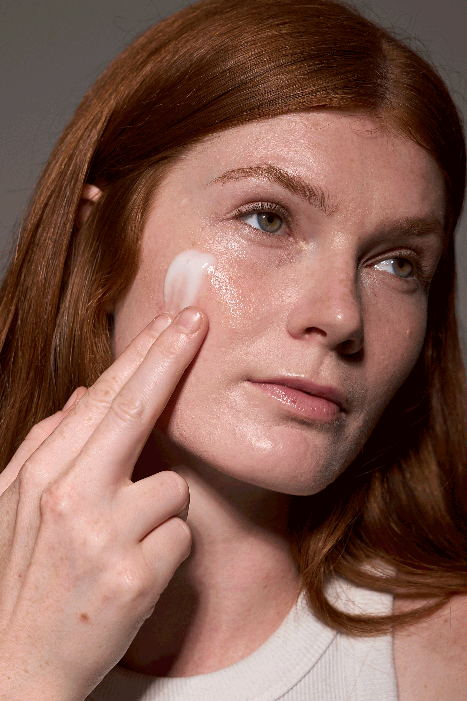 A woman with red hair applies white face cream to her cheek with her finger against a gray background.