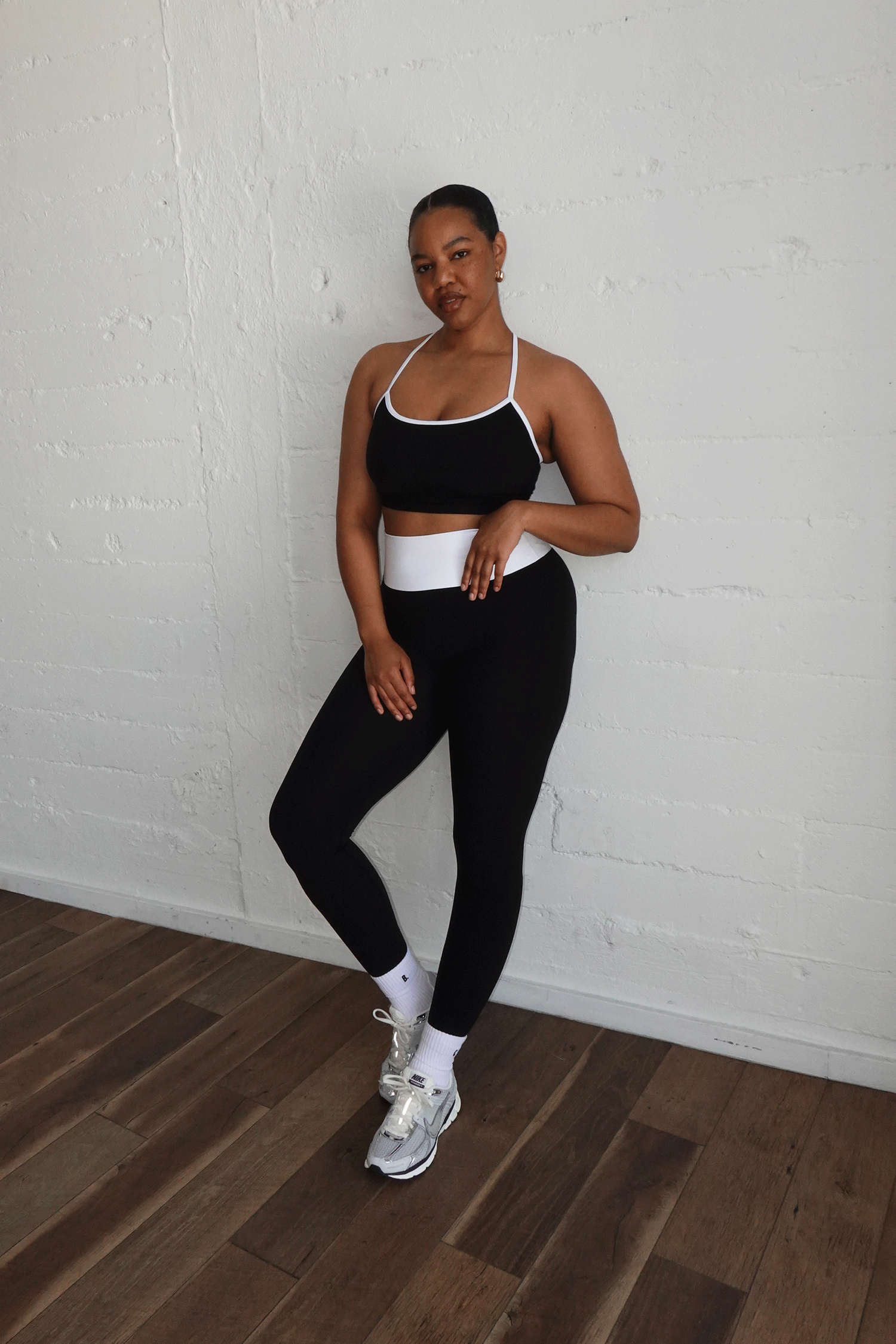 Woman in black sports bra and leggings with white accents poses against white brick wall in sneakers.