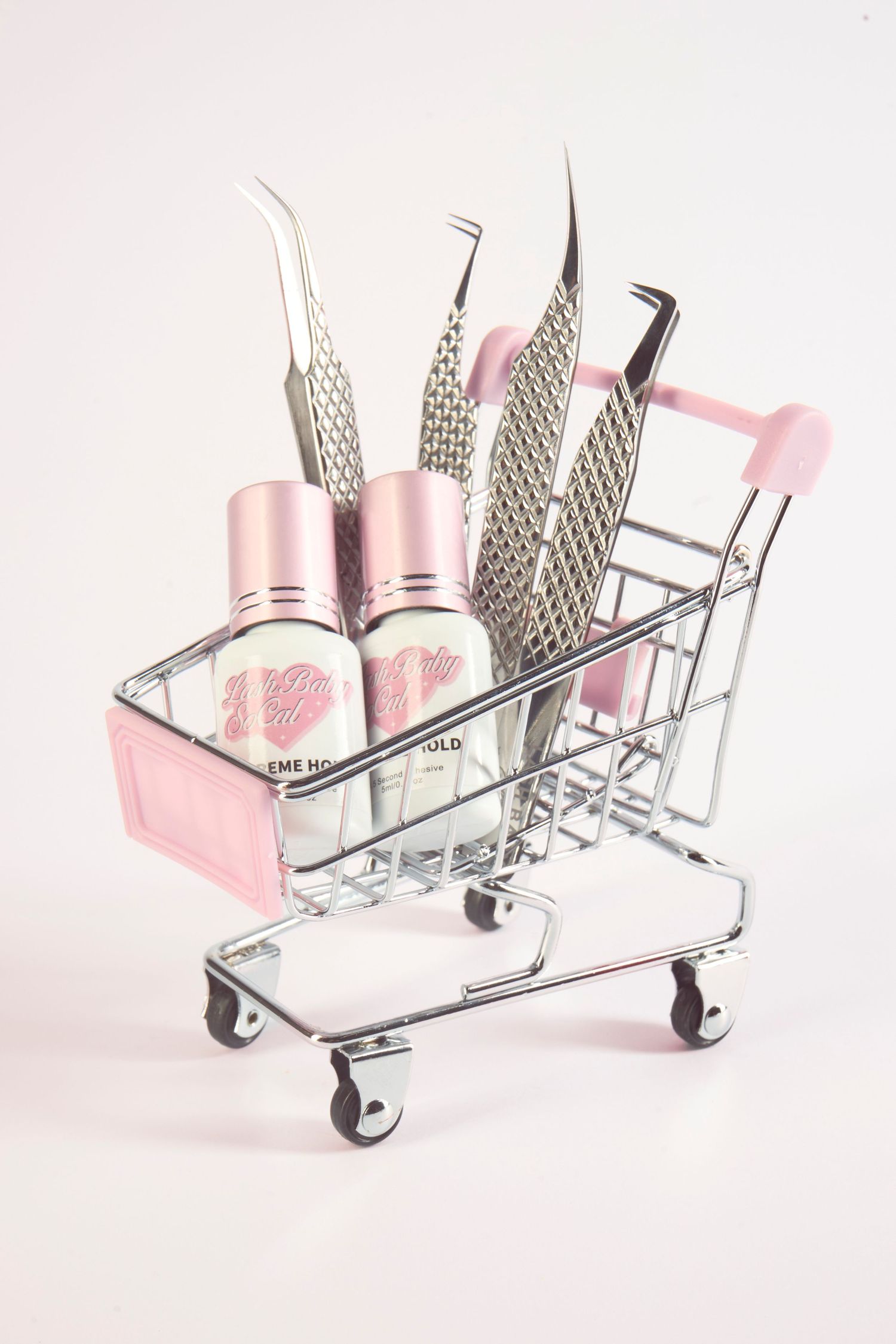 Pink mini shopping cart filled with tweezers and pink beauty bottles on a pink background.