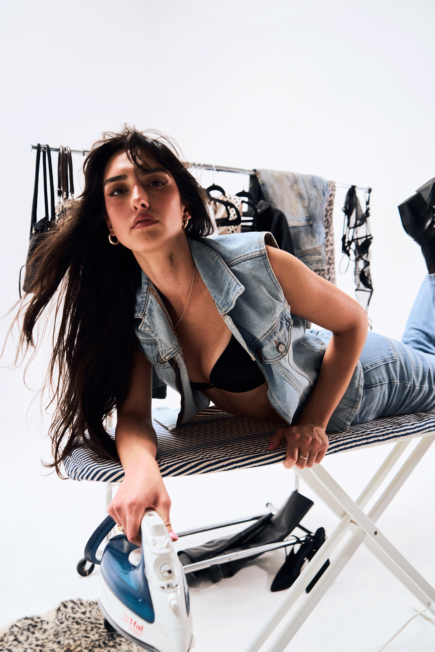 Young woman in denim vest and jeans poses on ironing board holding a steam iron in a studio setting.