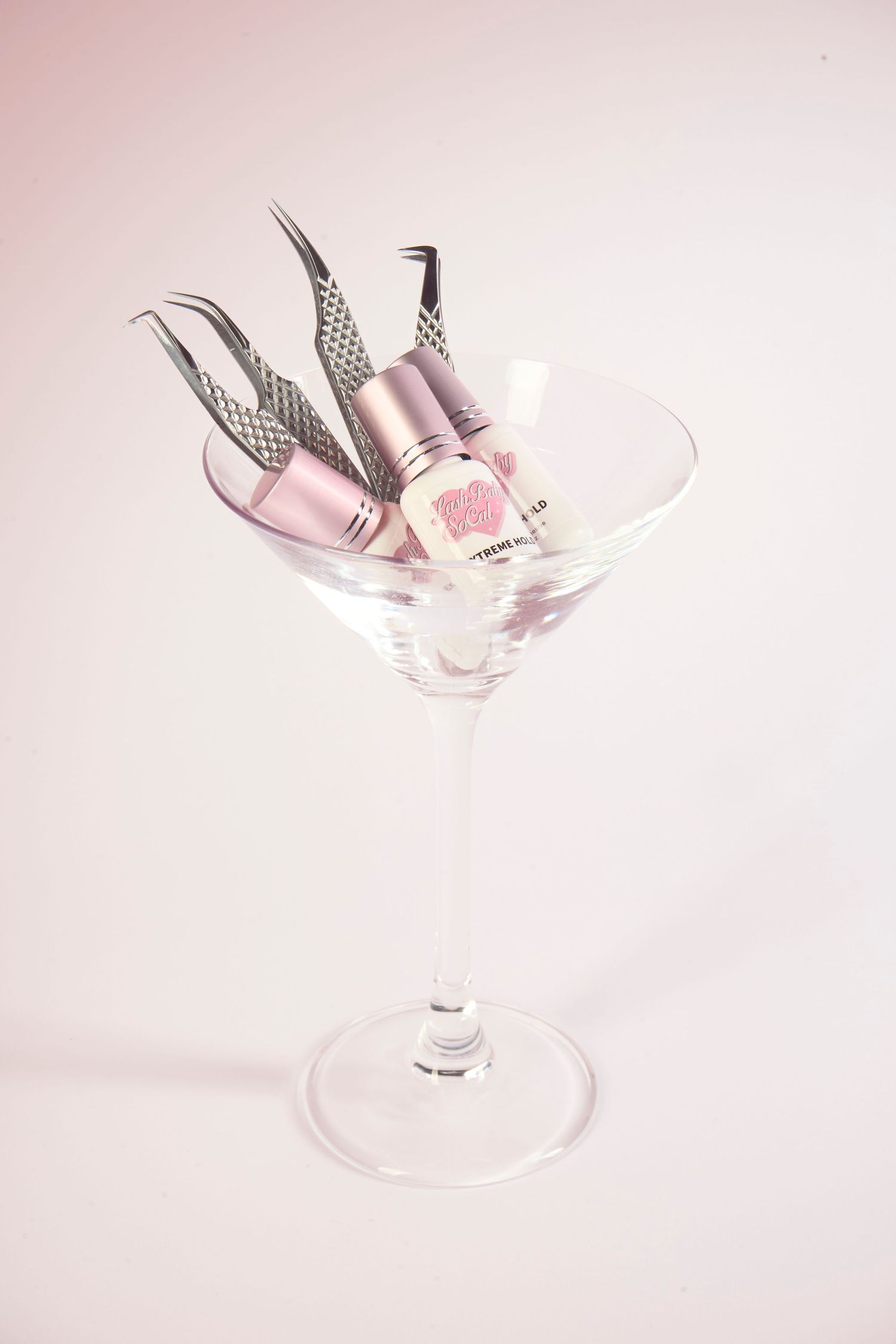 Pink nail polish bottles and tweezers displayed in a martini glass on a light pink background.