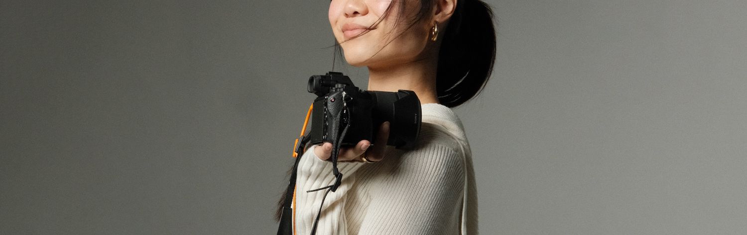 Smiling Asian woman holding a DSLR camer on her shoulder against a gray background.