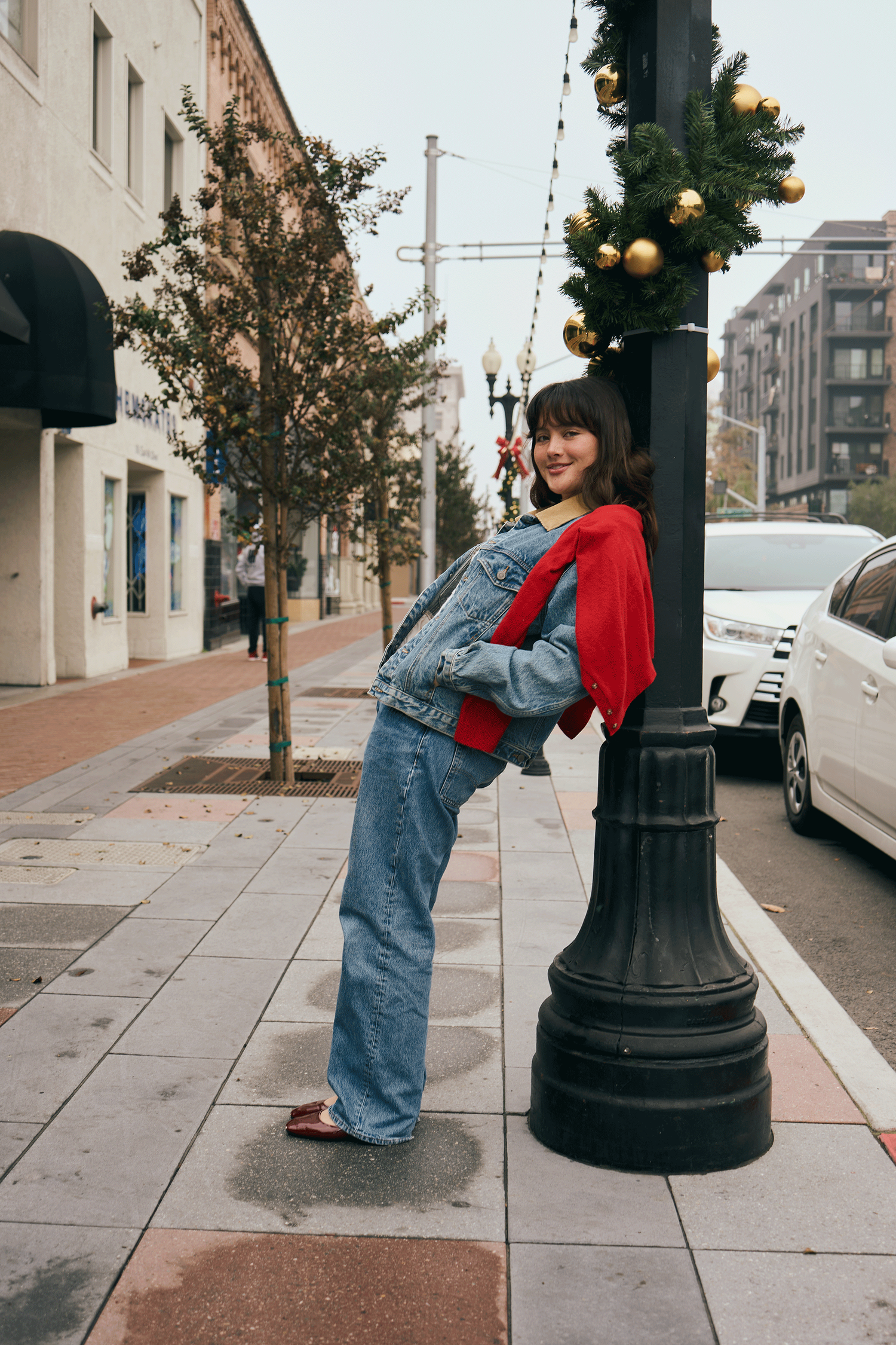 A smiling woman in a denim outfit leans against a Christmas-decorated lamppost on a festive urban sidewalk.