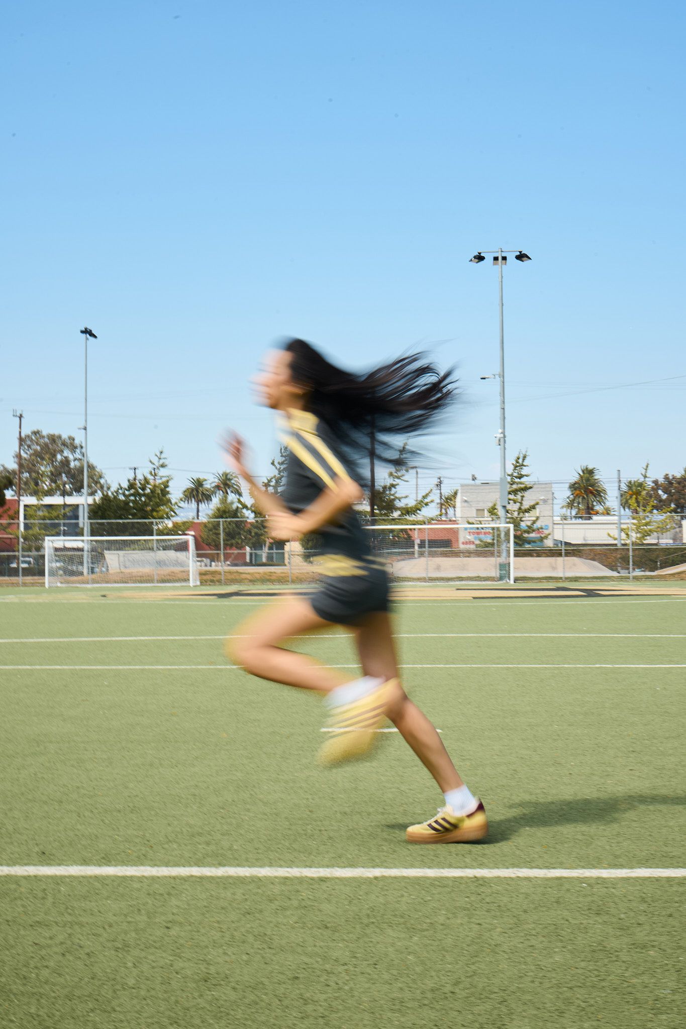 Female athlete sprinting across a green sports field, hair flowing, wearing gold and black uniform and cleats.