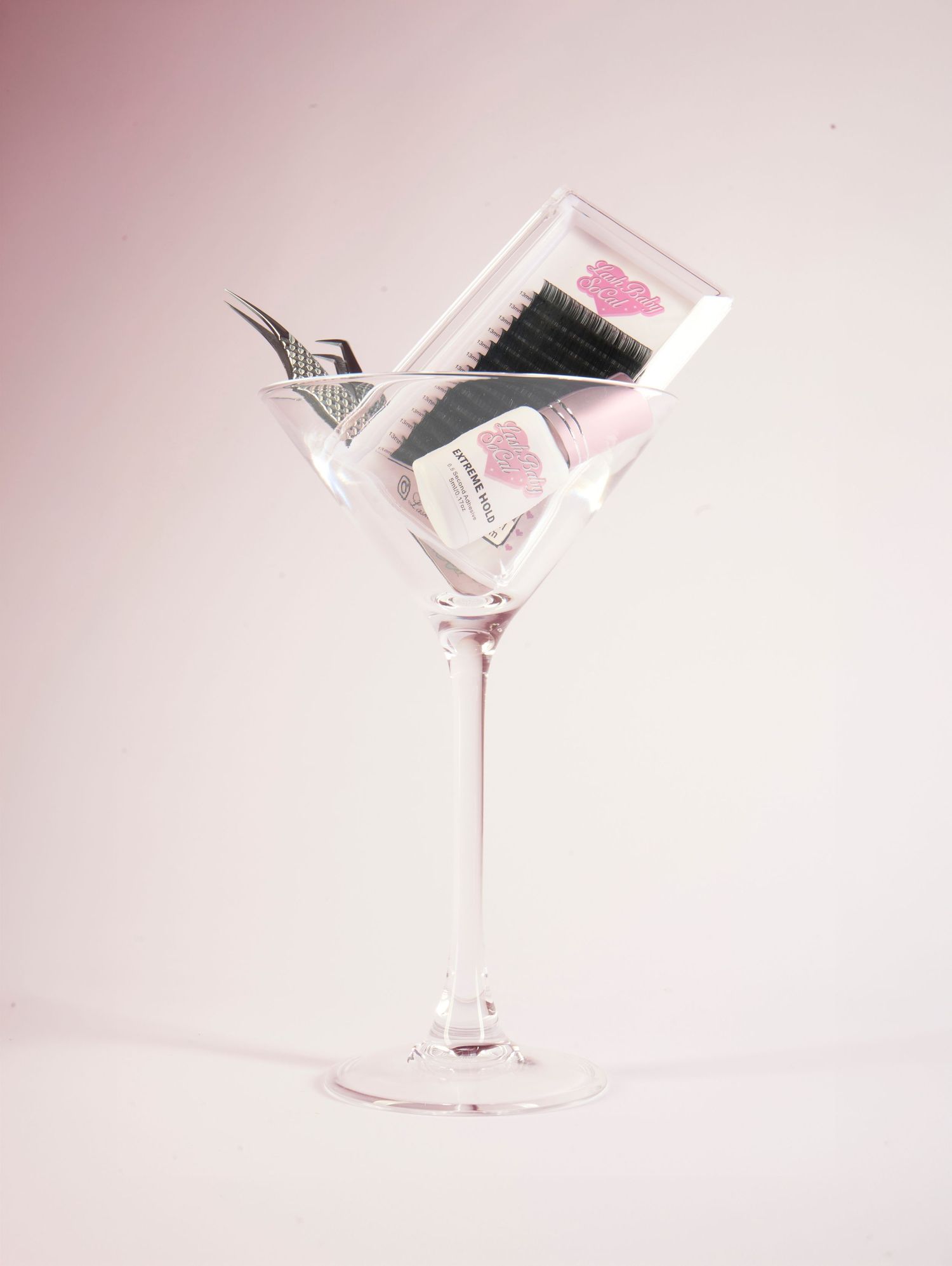 A martini glass filled with beauty tools including a hair brush and cosmetic products on a pink background.