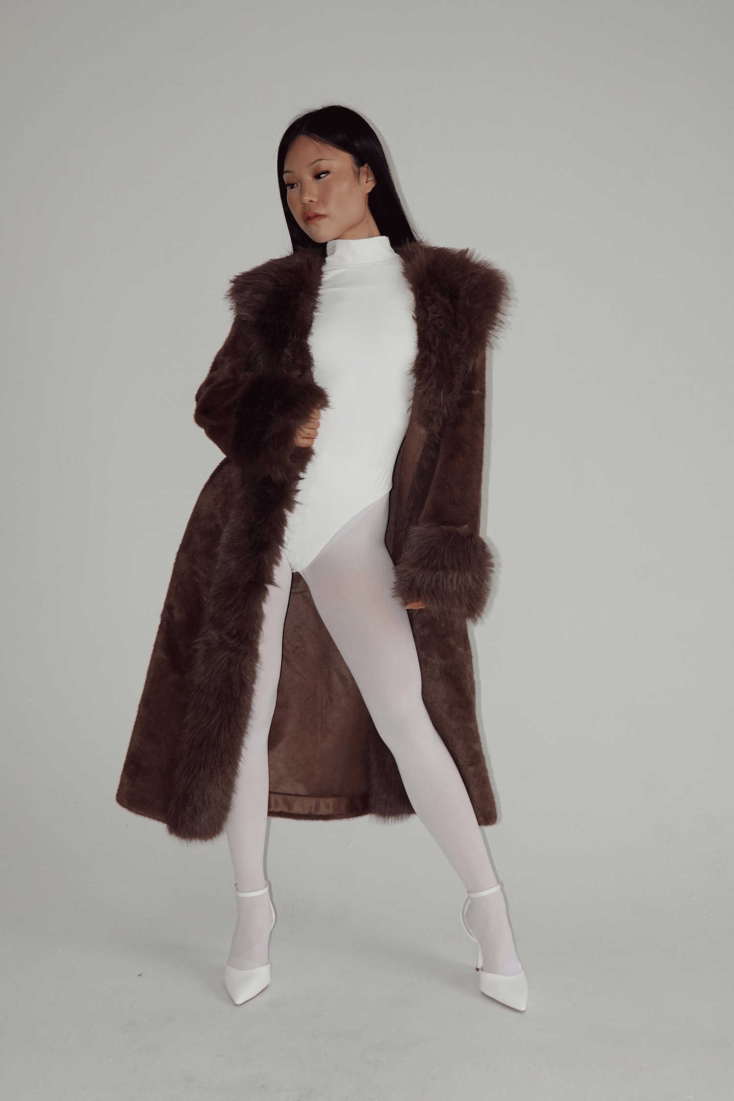 Woman wearing a long brown faux fur coat over a white turtleneck bodysuit with white tights and heels.
