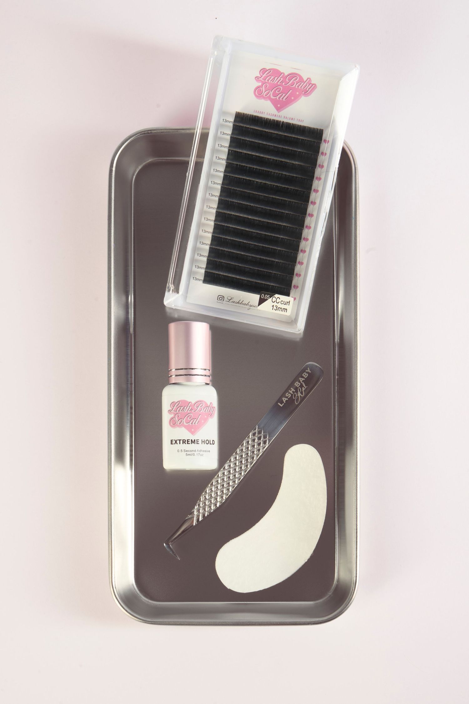 Eyelash extension kit with lash tray, extreme hold glue, tweezers, and under-eye pad on a metal tray.
