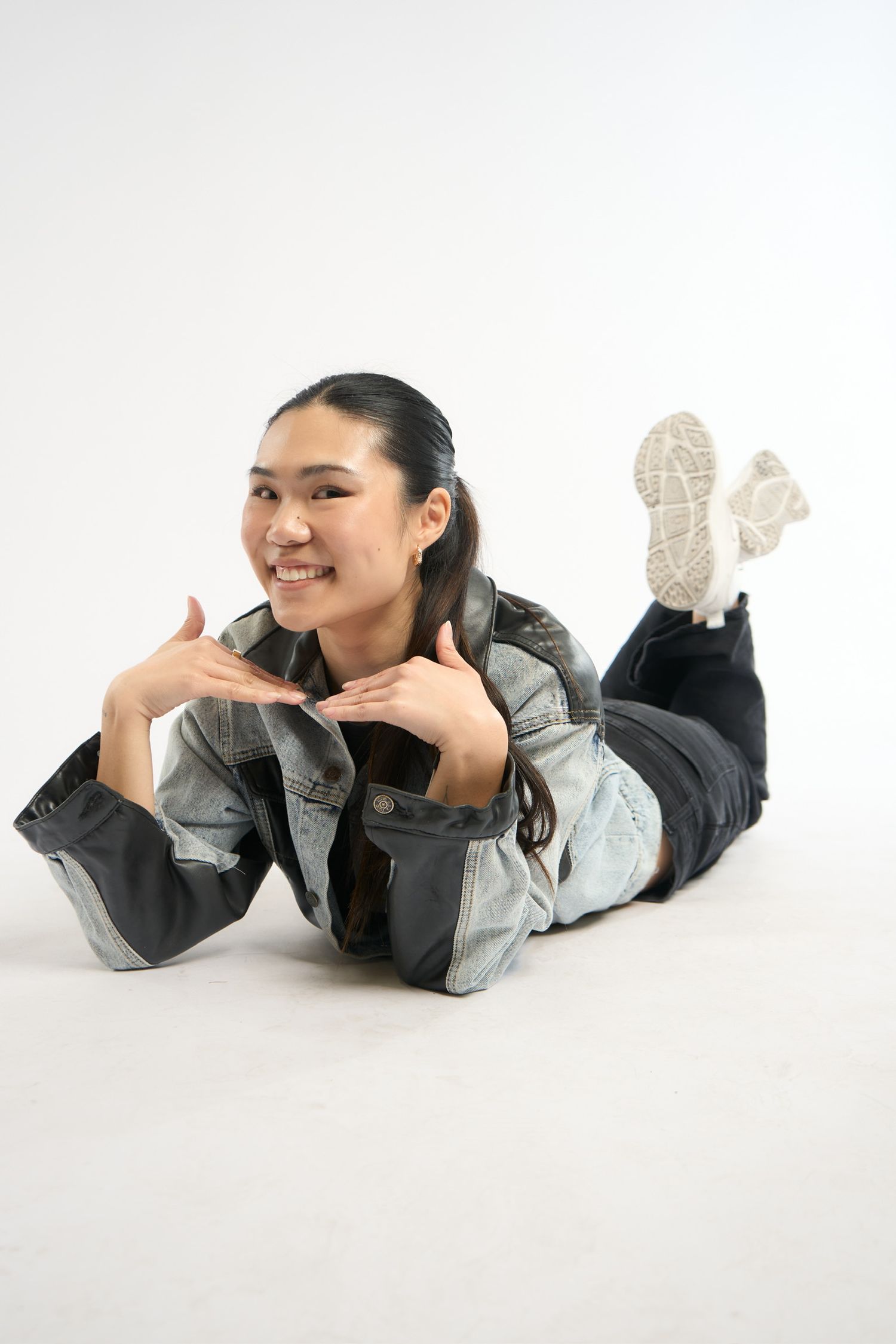 Smiling young Asian woman lying on floor in denim jacket and black jeans, posing with hands framing face.