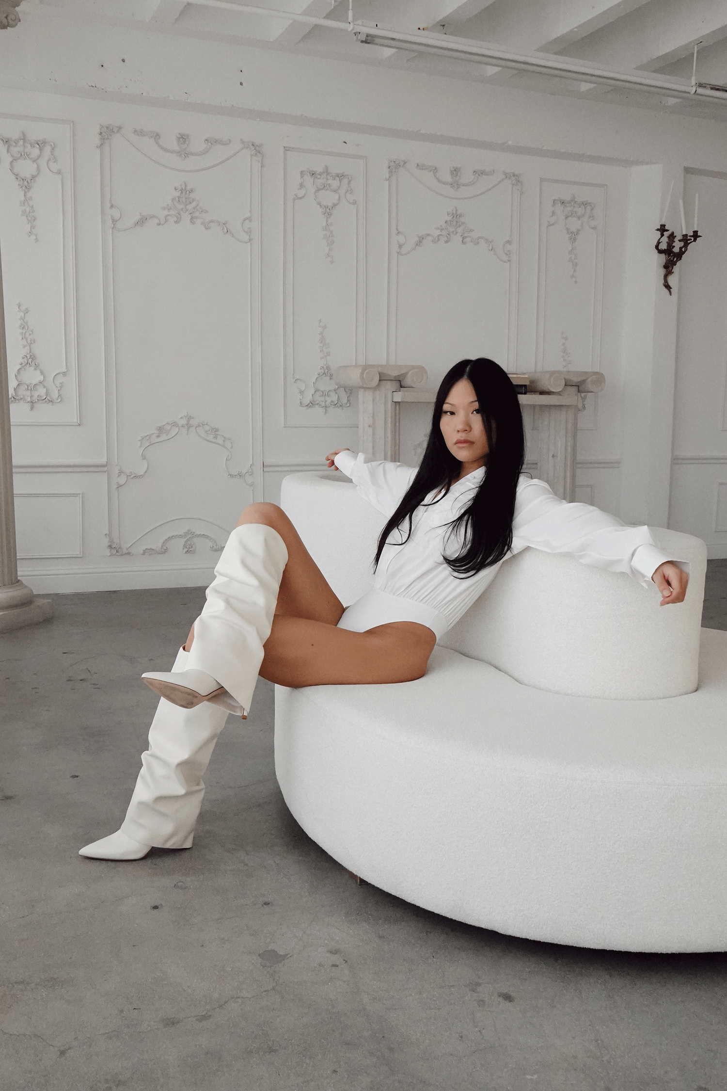 Woman in white bodysuit and knee-high boots poses on a curved white sofa in an elegant white room.