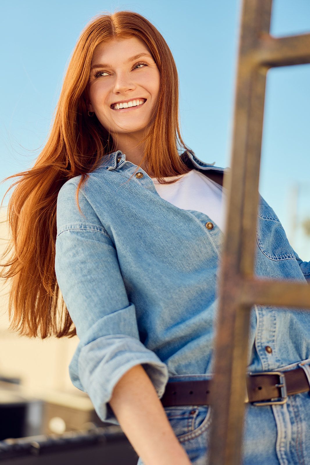 Smiling young woman with long red hair wearing a denim shirt and belt, posing outdoors against a blue sky.