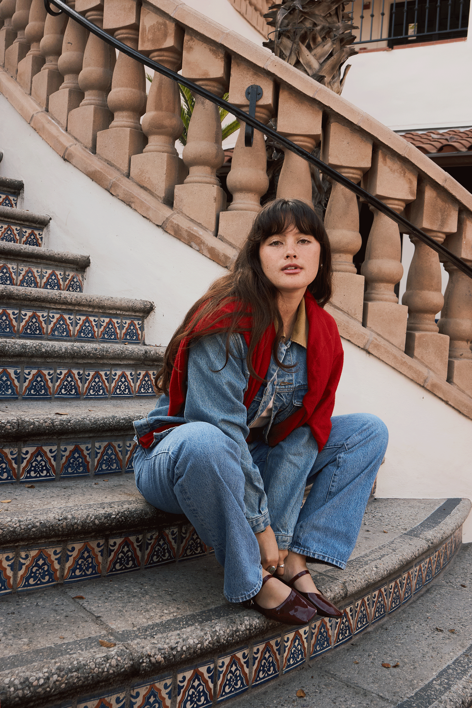 Woman in red cardigan and denim outfit sitting on ornate tiled stairs with stone balustrade.