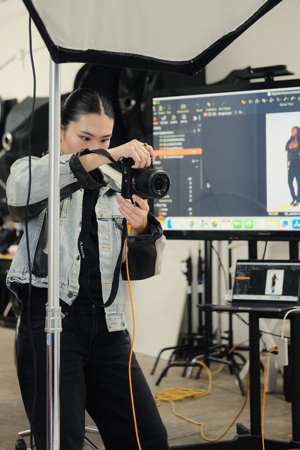 Female photographer in denim jacket aims DSLR camera in professional studio with lighting equipment and monitors.