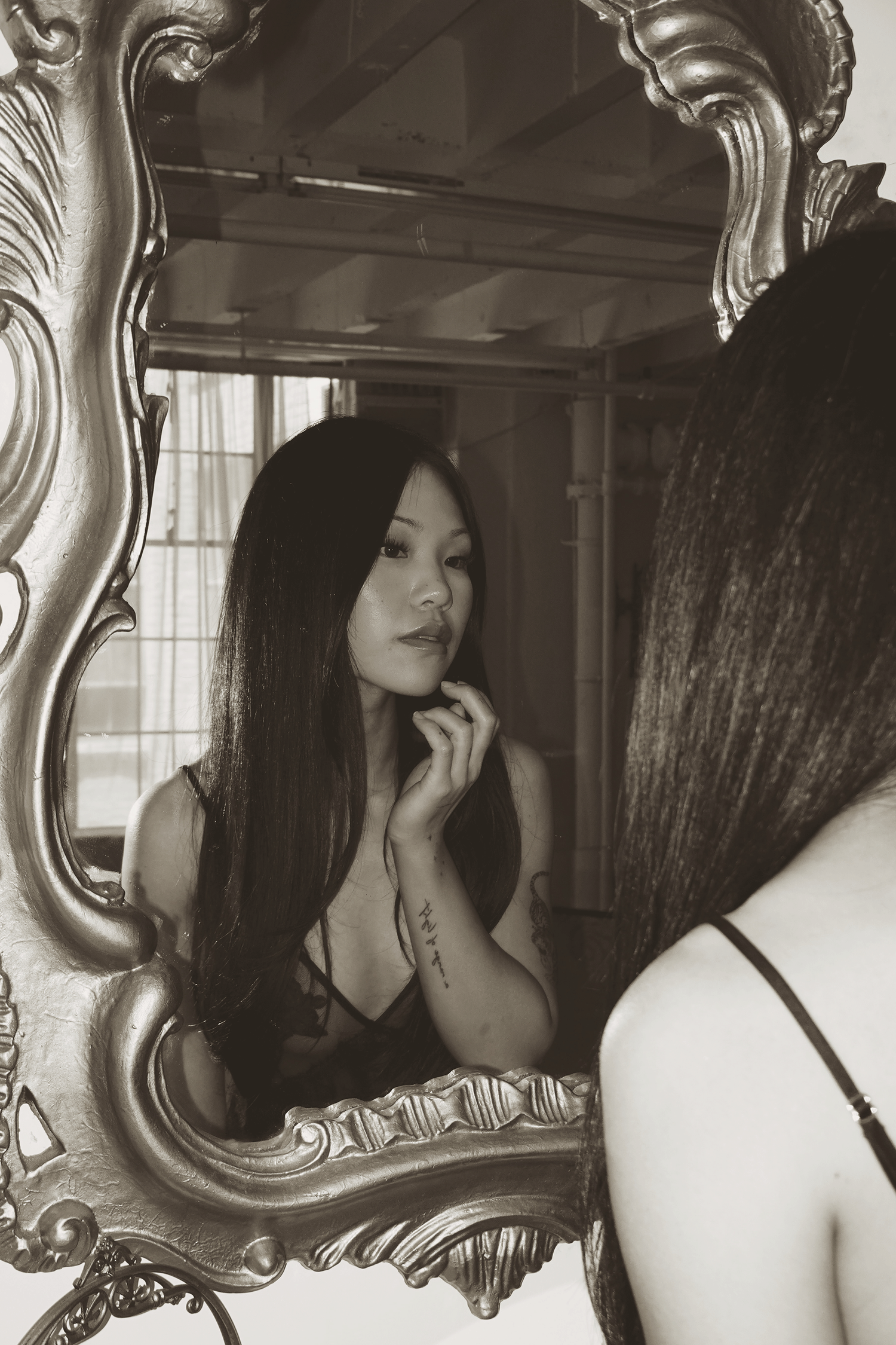 A woman with long dark hair gazes at her reflection in an ornate gold mirror in a sepia-toned portrait.