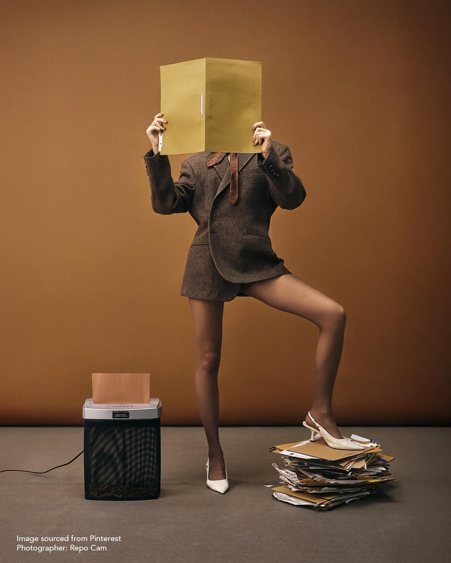 Person in brown suit balancing on one leg, holding a box over their face near a paper shredder and stack of files.