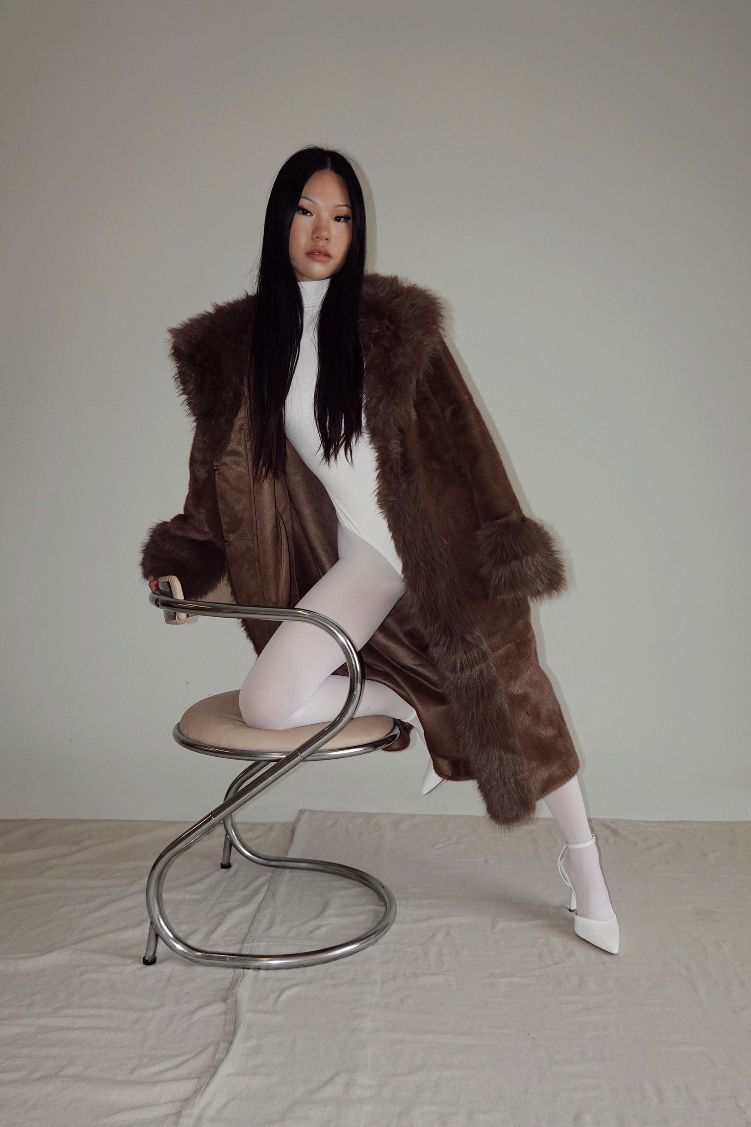 Woman in brown fur coat and white turtleneck bodysuit with white boots poses on chrome chair.
