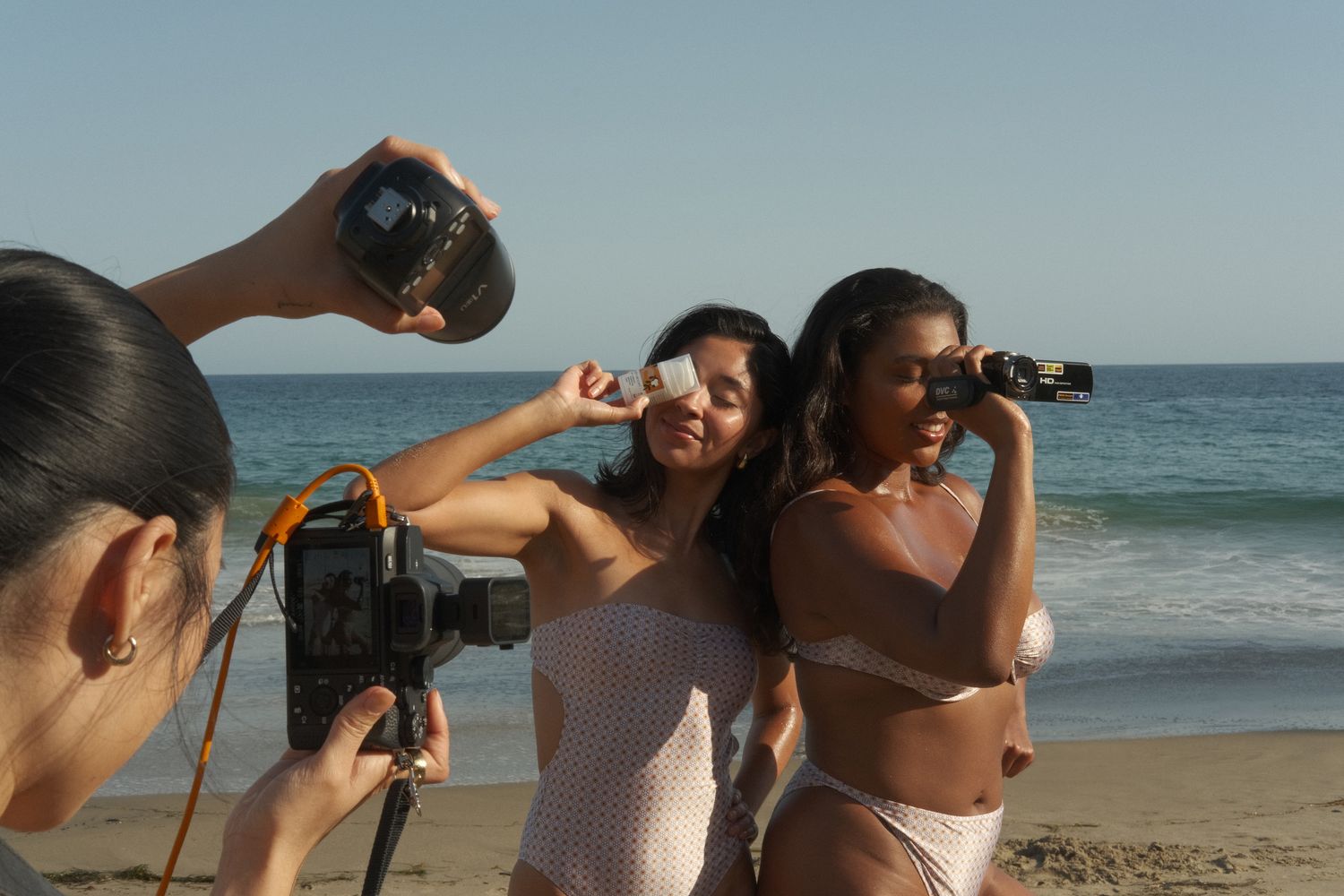 Two women in bikinis pose on a beach while a photographer captures their photo with a professional camera.